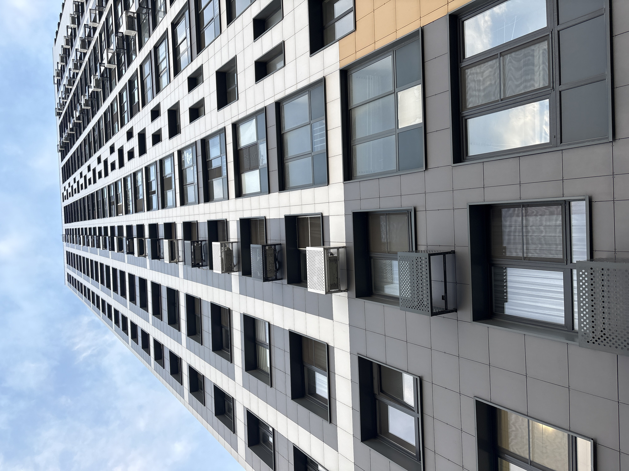 A low-angle, wide shot captures the facade of a modern, tall apartment building under a cloudy sky. The building, with its many windows and sleek, tiled exterior, is angled upwards and to the left, dominating the frame. The facade is characterized by a repeating pattern of white and grey tiles, interspersed with dark-framed windows. Some windows appear to be open, with what look like air conditioning units or ventilation systems visible in the openings, protected by perforated metal screens.
The building's design is contemporary and geometric, with a sense of verticality emphasized by the upward perspective. The sky is a soft blue with scattered white clouds, suggesting a daytime scene, possibly overcast or with the sun behind the clouds. There are no people visible in the image. The overall impression is one of urban architecture, highlighting the structured and modern design of the residential building. The location context indicates the building is in Лесные Поляны, Russia, though no specific landmarks are visible to confirm this. No text is present in the image.