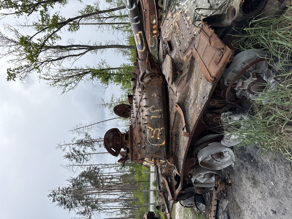 A burnt-out, heavily damaged Russian T-72 tank sits abandoned in a wooded area in the town of Dmytrivka, Ukraine, located at coordinates (latitude: 50.464, longitude: 30.182). The tank is facing right, partially obscured by foliage and trees. Its turret is missing, and the hull is covered in rust, dirt, and debris. Multiple sections of the track are missing, and the tires are shredded or completely gone. The tank appears to have been struck by multiple rounds of ordnance. Numerous bullet holes and scorch marks are evident on the hull, turret, and gun barrel. The image was likely taken sometime after the initial battle. The tank is surrounded by green vegetation and stands in the middle of a road that is visible in the background.