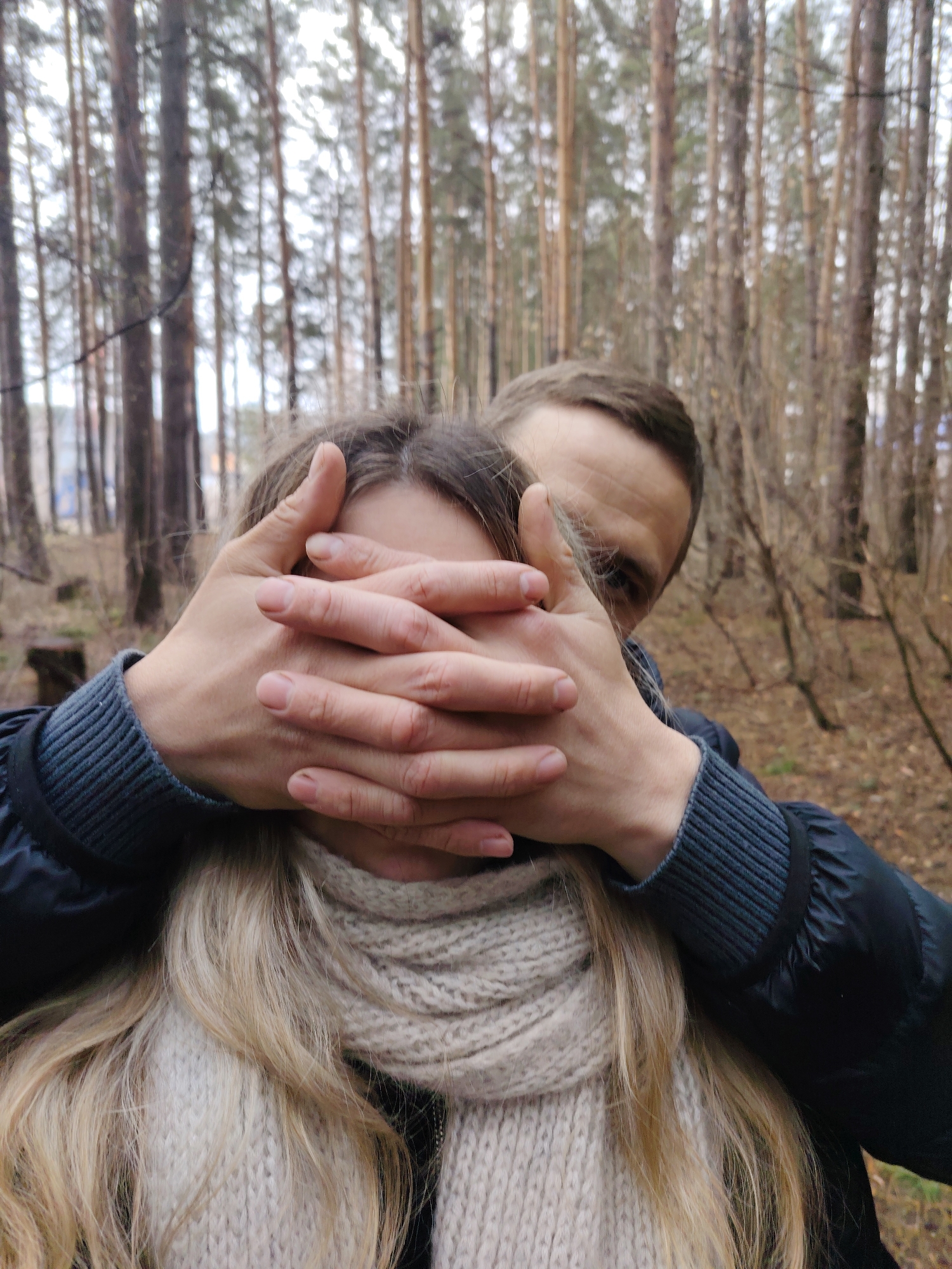 The image features two individuals in a forest, likely in Chaikovsky, Russia. A male figure is shown from the chest up, with his hands covering the mouth and lower face of a female companion. His eyes are visible above his hands, looking directly at the camera with a subtle, possibly playful, expression. He wears a dark blue or black jacket with a ribbed cuff visible on his left arm. The female's blonde or light brown hair cascades over her shoulders, and she wears a cream-colored, thick-knit scarf. Her face is entirely obscured by the male's hands.
The setting is a dense forest, characterized by numerous tall, slender tree trunks that stretch into the background. The ground is covered with dry foliage and sparse undergrowth, suggesting an autumn or early spring season. The lighting is soft and diffused, indicating an overcast sky or twilight conditions, with no strong shadows. No text is visible in the image.
