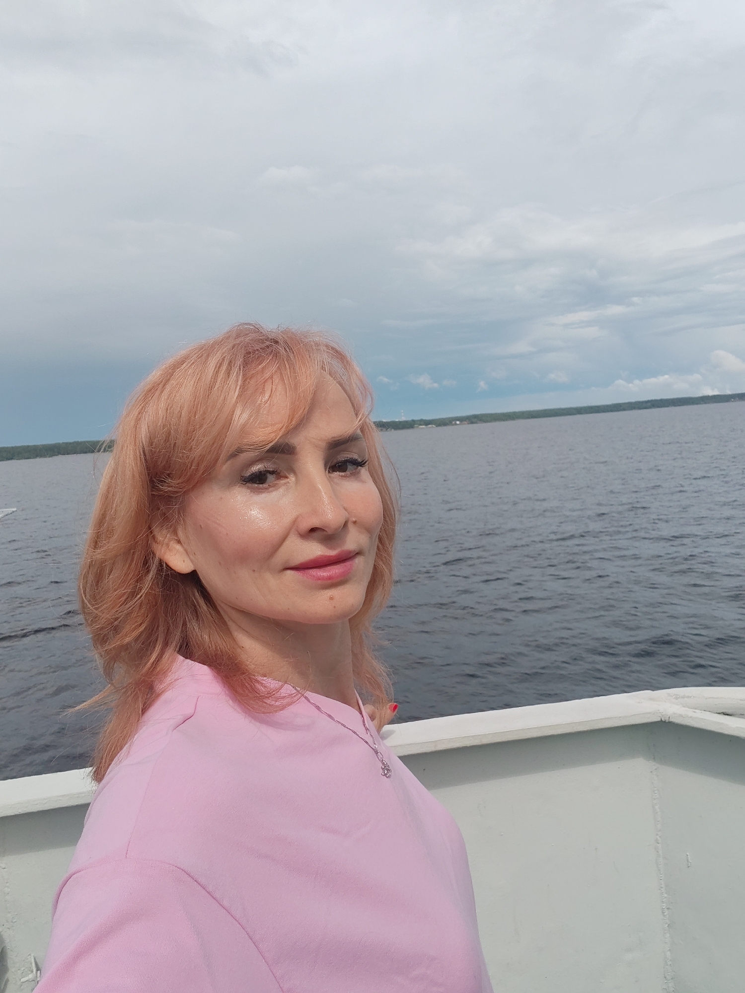 A woman with blonde hair, wearing a pink shirt, is standing on a white boat deck. She is looking at the camera with a slight smile. The water is dark and calm, with a distant shoreline covered in trees. The sky is overcast with scattered clouds. The location is identified as Kostyaevka, Russia.