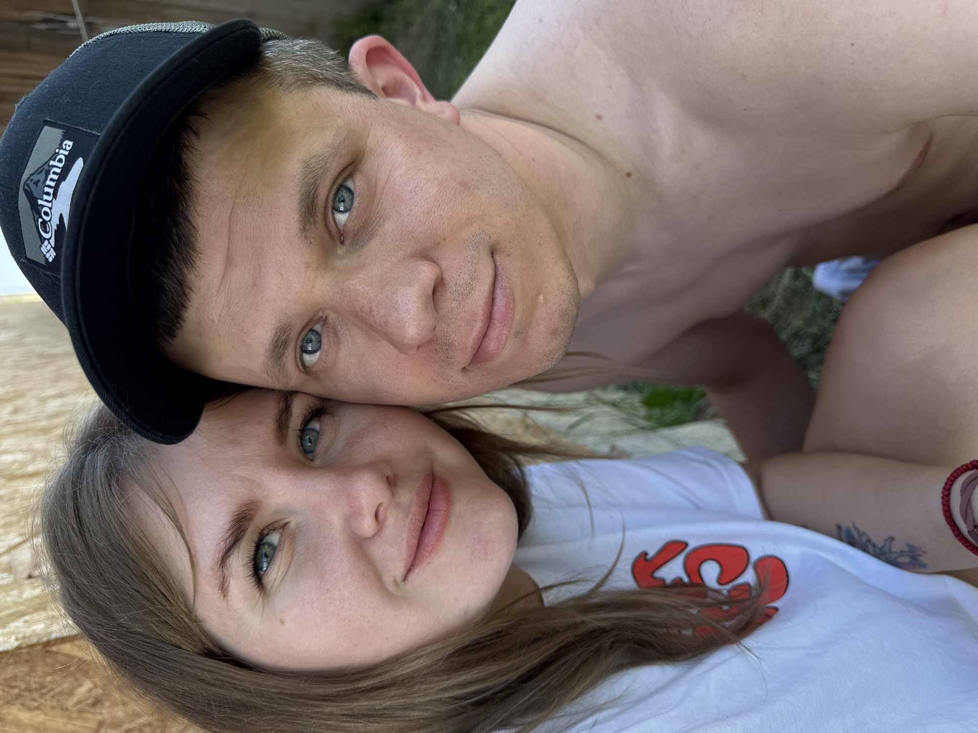 A close-up shot depicts a man and woman lying together. The man is wearing a black Columbia baseball cap. He has light skin, blue eyes, and short, light-colored hair. He is shirtless, and a scar is visible on his upper chest. The woman has light skin, blue eyes, and shoulder-length brown hair. She is wearing a white t-shirt with a red design on it. A red bracelet is visible on her left wrist, along with a tattoo on her forearm. They appear to be outside, with grass and what seems to be wood visible in the background. No information indicates this image was taken in Фоки, Russia.