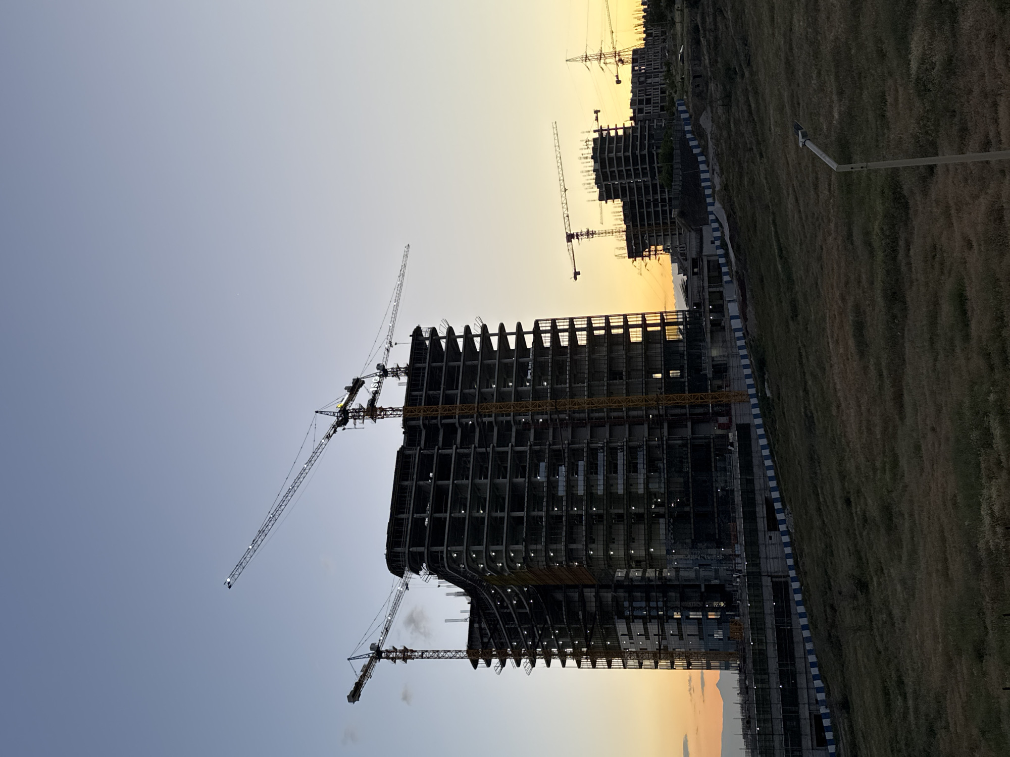 This media file captures a large-scale construction site in Yerevan, Armenia, during the soft light of either dawn or dusk. The image, oriented vertically, prominently features several high-rise buildings in various stages of construction.
In the foreground, a tall, multi-story building with a distinctive curved facade dominates the left side of the frame. Its concrete or steel skeleton is clearly visible, with some lower floors already fitted with windows, from which artificial lights glow, suggesting the time is either early morning or evening. Multiple tower cranes are positioned around and atop this structure, their booms extending across the sky. Further in the background, more unfinished buildings and cranes are discernible, indicating a vast development project.
To the right of the construction zone, a wide expanse of dry, tan-colored grass stretches into the distance, appearing as an undeveloped area adjacent to the construction. A single street lamp stands within this grassy field. A barrier, striped in blue and white, separates the construction site from the open land.
The sky transitions from a warm, soft yellow-orange near the horizon to a pale, clear blue-grey overhead, confirming the twilight hours. No clouds are visible, and the weather appears clear. There are no people visible in the image, and no discernible text. The scene conveys a sense of ongoing urban development and expansion.