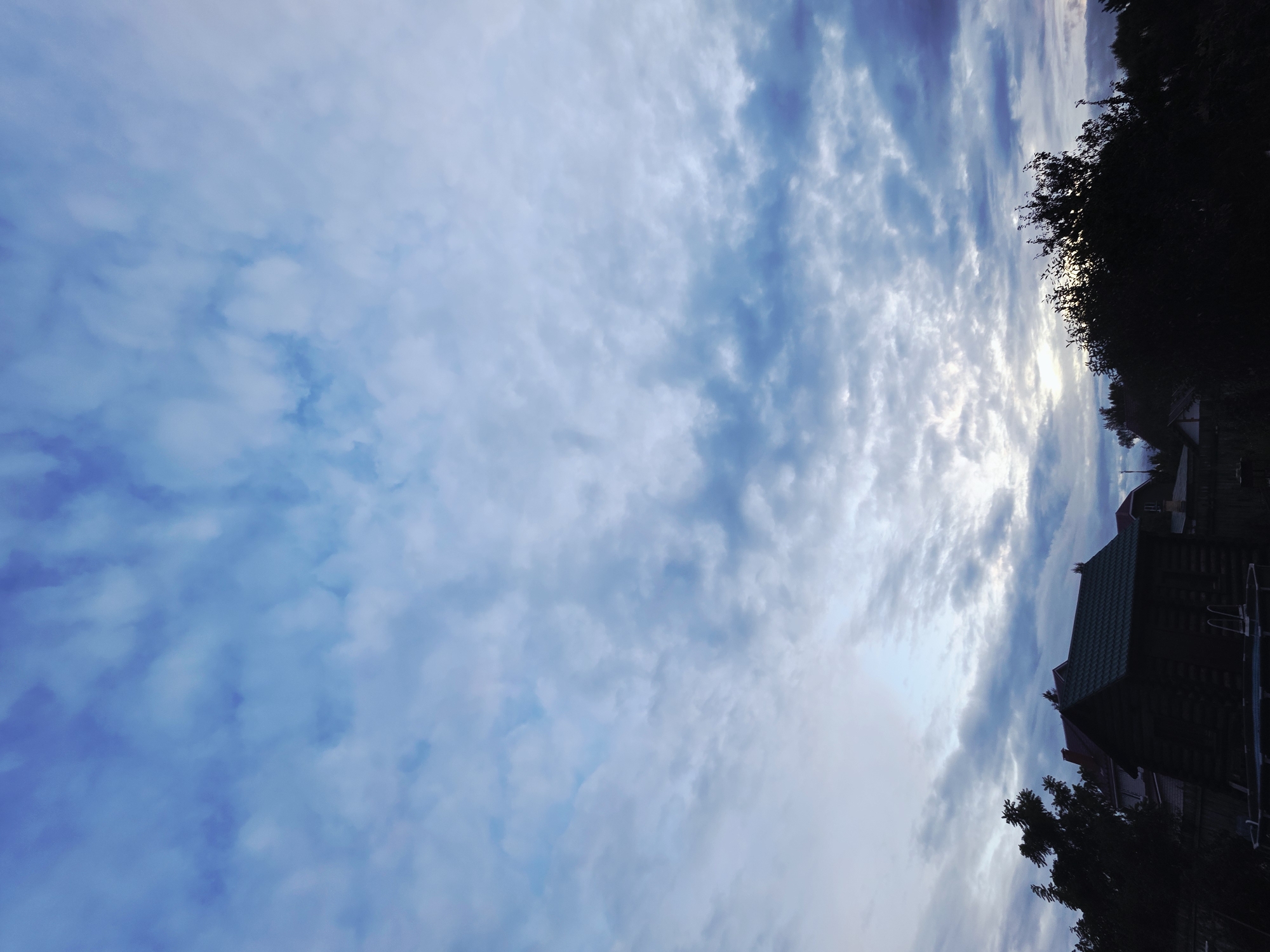 The image depicts a cloudy sky over a residential area in the city of Rechitsy, Russia. Silhouetted against the sky are the dark forms of trees and buildings, including what appears to be a wooden house with a green roof and a fence. A small round swimming pool with a ladder is partially visible in the foreground. The sky is filled with scattered clouds, with patches of lighter sky suggesting the presence of the sun behind them, possibly indicating dawn or dusk. The overall mood is subdued, with the low light casting the foreground elements into shadow.