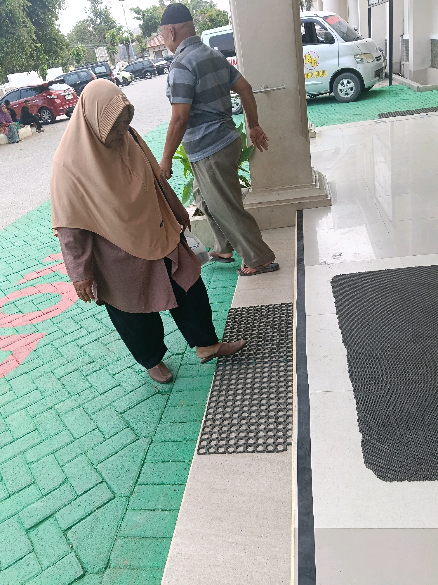 In Balun, Indonesia, an elderly woman wearing a tan hijab and a brown tunic walks on a green tiled path. To her right, a man in a grey striped shirt and olive green pants walks towards the foreground, looking back at her. A white van with "AE" and "UNG PEDULI" written on it is parked in the background. The scene appears to be an outdoor walkway near a building.