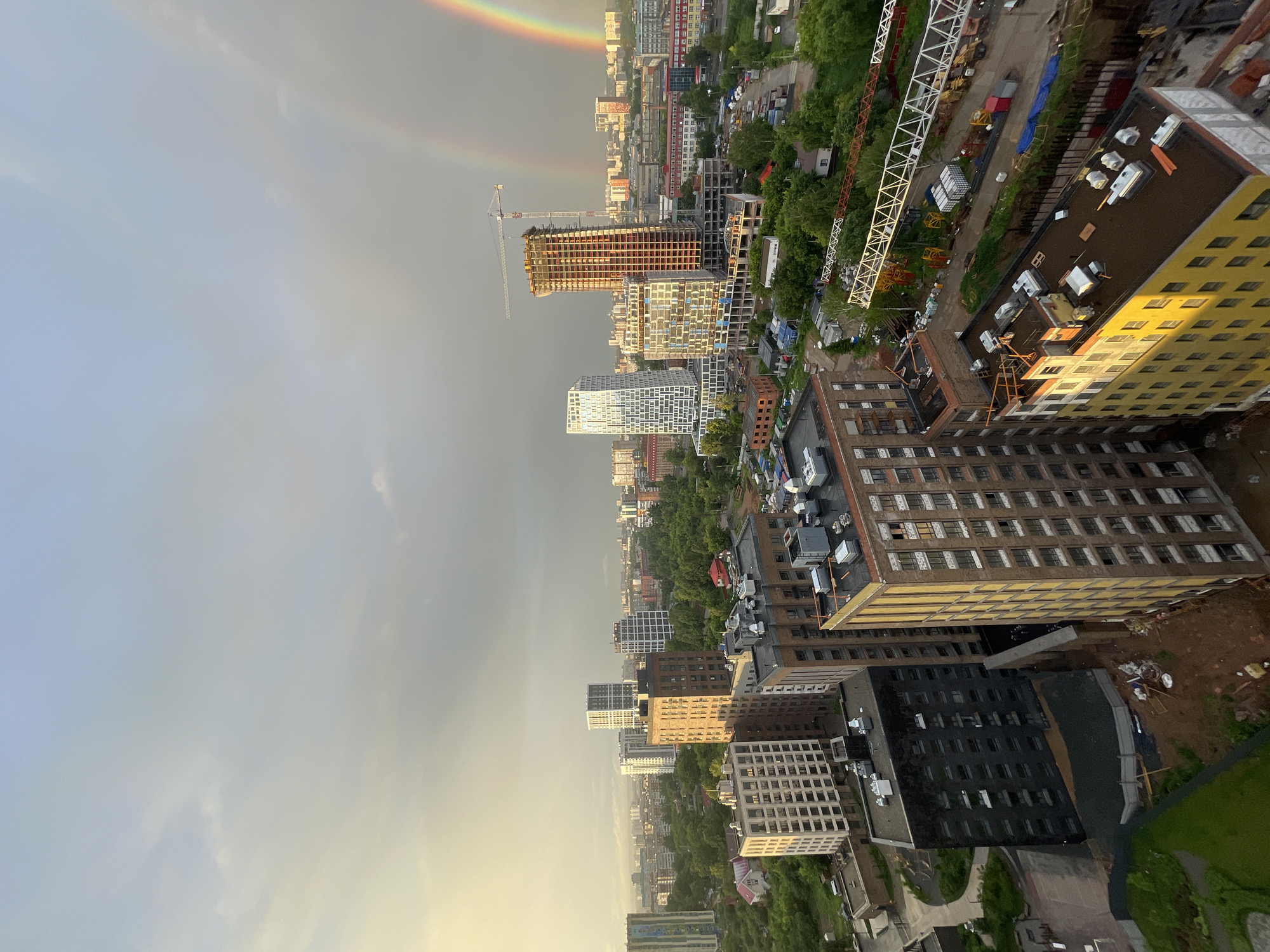 This is a high-angle, wide shot of a cityscape at dusk. The sky is overcast with a soft, golden light on the left side, indicating the setting sun. A double rainbow arcs across the sky on the right side of the frame, with both the primary and secondary bows clearly visible. The cityscape is densely packed with buildings of varying heights and architectural styles, interspersed with lush green trees.
In the foreground, several apartment buildings are prominent. One building on the right is a tall, yellow brick structure with a dark brown roof, showing signs of ongoing construction with exposed beams and scaffolding. To its left is a dark grey, rectangular building. A lighter beige, modern apartment building with numerous windows is situated in the middle left. Behind these, more buildings stretch into the distance, including a few under construction with cranes visible.
The scene appears to be a developing urban area, with a mix of completed residential buildings, some older structures, and active construction sites. The presence of trees suggests a relatively green city or a park within the urban environment. No people are visible in the frame. The overall atmosphere is serene, illuminated by the natural light of the setting sun and the dramatic appearance of the double rainbow.