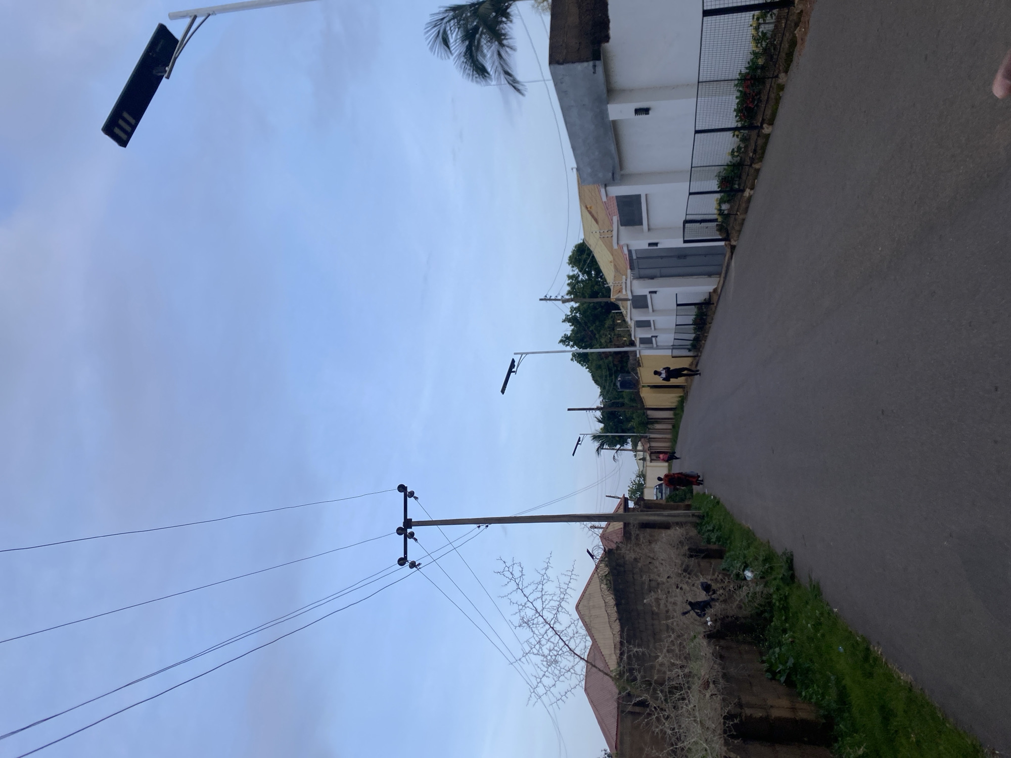The image depicts a street scene in Barikin Jirga Summa, Nigeria. The street is paved and runs towards the background. On the left side of the street, there is a low, rough brick wall with a dry, thorny shrub growing against it. Behind the wall are houses with red roofs. Further down the street on the left, there are more houses and a utility pole with multiple wires extending upwards. On the right side of the street, there are modern white walls with fences in front of them, some with flowering plants. Several streetlights, including solar-powered ones, are visible along the street. A few individuals are walking on the street in the distance, two women in colorful clothing and two men in darker attire. The sky is overcast with a pale blue hue. The overall environment suggests a residential area with a mix of older and newer constructions.