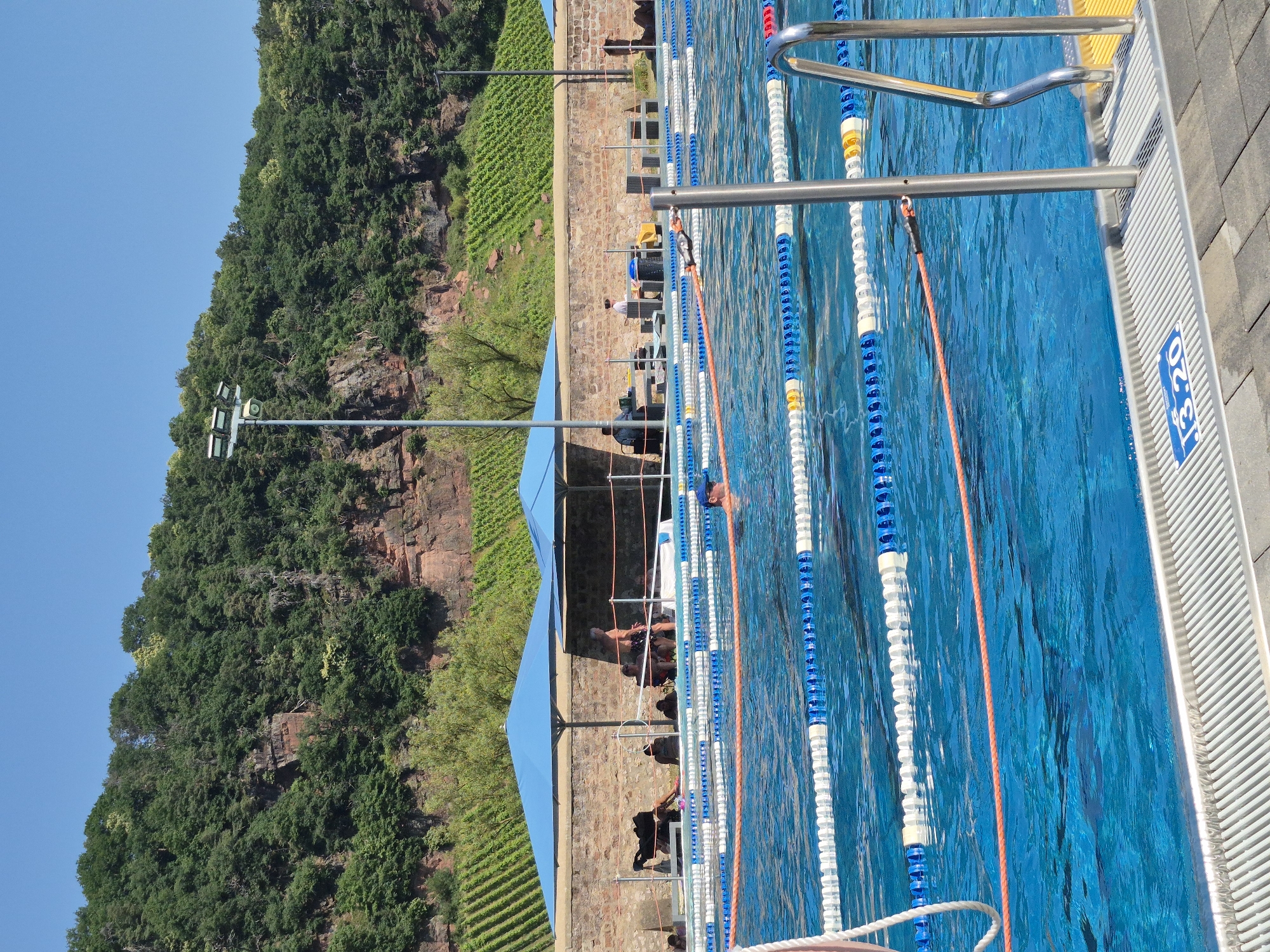 An outdoor swimming pool is shown with lane markers and a ladder. The pool is surrounded by a paved area and a stone wall. Beyond the wall, there is a steep, wooded hill with terraced vineyards. Several people are visible, some swimming in the pool and others sitting in the shaded area under blue umbrellas. The sky is clear and blue.
