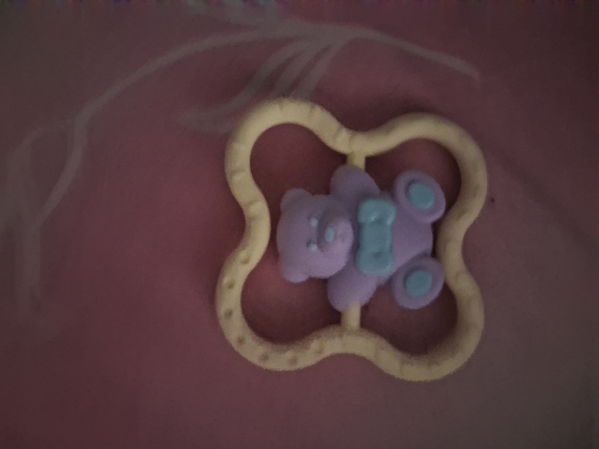 A close-up, slightly blurry image shows a pale yellow, heart-shaped rattle with a small, light purple teddy bear figurine in the center. The teddy bear has blue accents on its bow and feet. The background is a dark pink, textured surface. There is no indication of the city of Ivky, Ukraine in the image.