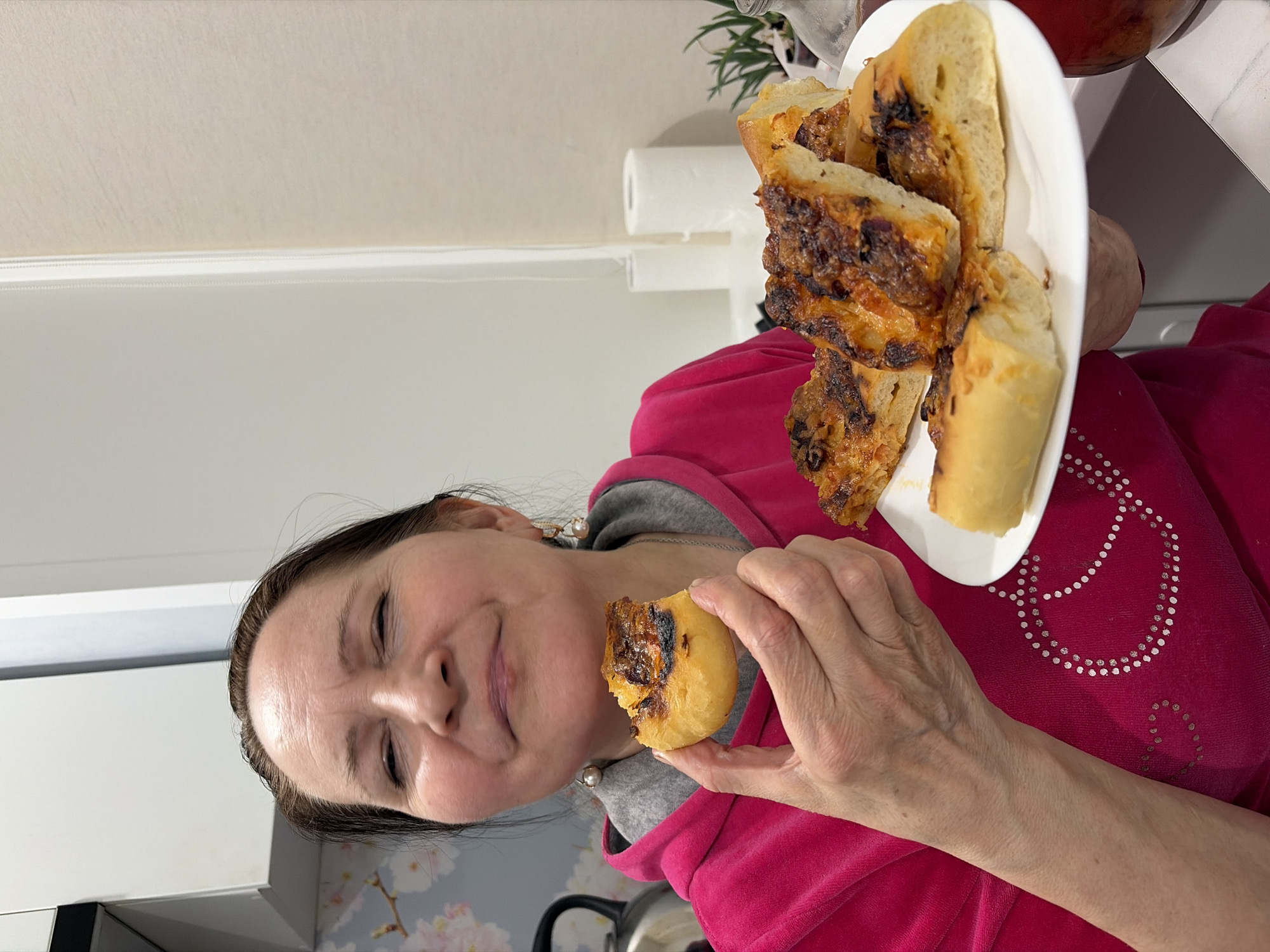 A middle-aged woman is indoors, holding a white plate filled with several pieces of what appears to be toasted bread topped with melted cheese and a meat mixture. She holds a single smaller piece of the same bread in her other hand, seemingly about to eat it. She is wearing a pink sweatshirt with a gray hoodie underneath, and pearl earrings are visible. The background includes white cabinets, wallpaper with a floral design, a roll of paper towels, and a glass container. The location is potentially in Neftekamsk, Russia, although this cannot be confirmed definitively from the image alone.