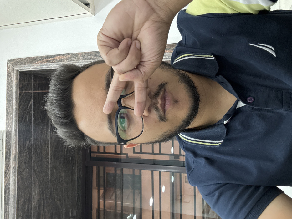 A close-up portrait shows a South Asian adult male with dark hair, a beard, and a mustache, wearing black-framed eyeglasses and a navy blue Nautica brand polo shirt with white and yellow striped collar details. He is looking directly forward, with his right hand raised to his face, forming a V-shape with his index and middle fingers, partially obscuring his right eye and the bridge of his nose. The background is an indoor setting, featuring a dark granite-patterned surface in the upper left, a window or glass partition with a grid pattern behind the subject through which blurred lights are visible, and a plain light-colored wall or cabinet section in the upper right. The specific location within the city of Kolkata, India, cannot be determined from the visual information provided in the image.