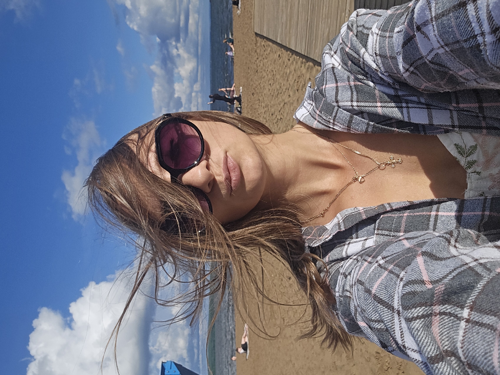A woman is taking a selfie on a sandy beach with a wooden boardwalk and the Kama River in the background. She is wearing oversized sunglasses, a plaid shirt over a white floral top, and a gold necklace with a cross pendant. Her hair is blowing in the wind. Several other people are visible in the background on the beach and in the water, some lying down and others standing. The sky is blue with scattered clouds. This scene is likely in Chaikovsky, Russia, a city known for its scenic riverbanks and recreational areas.