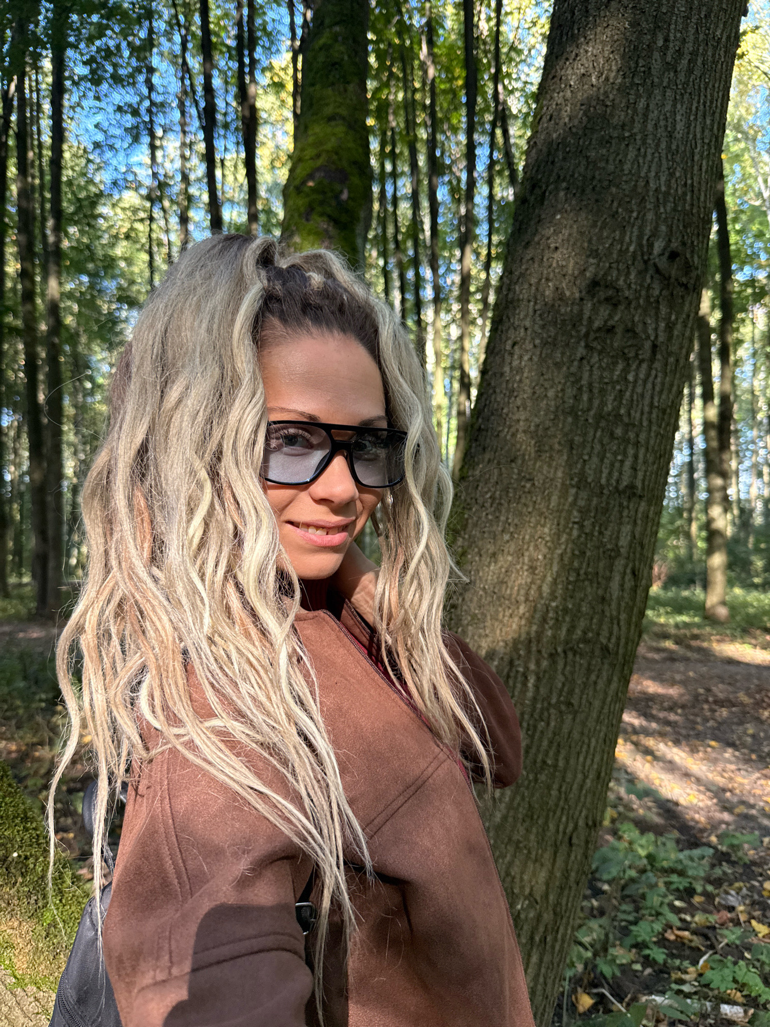 A close-up, eye-level, medium shot captures a woman with long, blonde, wavy hair and a brown jacket, wearing black sunglasses and a red top, smiling as she stands in a forest. The woman is positioned to the left of a large tree trunk, with a forest of tall trees and dappled sunlight visible in the background. The ground is covered with fallen leaves. The scene is set outdoors, with the natural environment of a forest. The woman is looking towards the camera. The image does not provide any information about the city of Rusanovo, Russia.