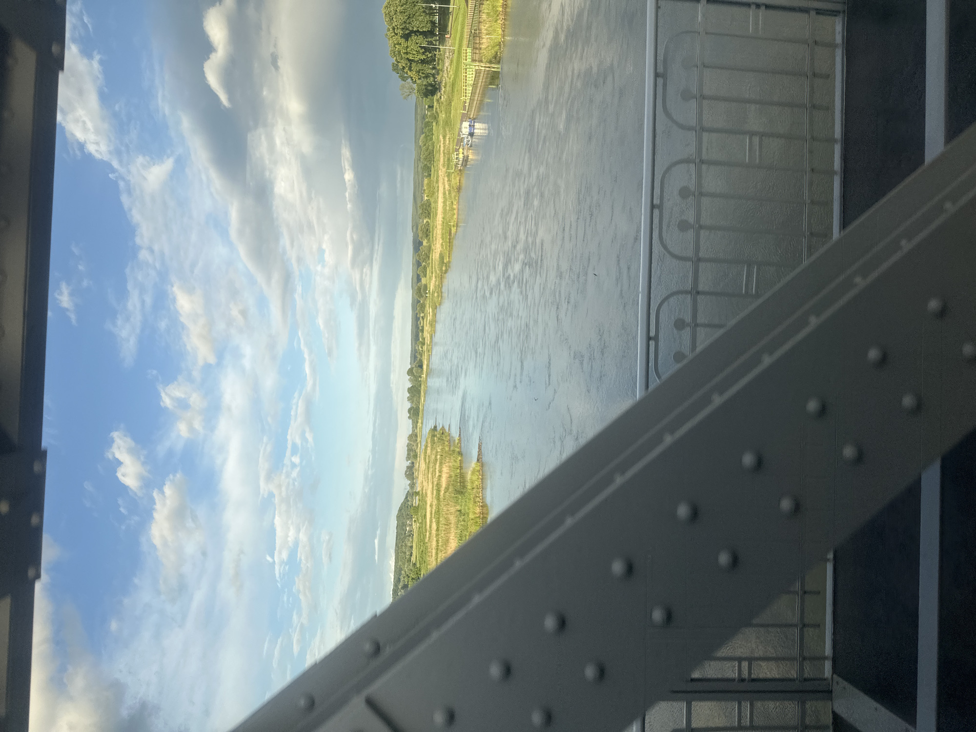 The image captures a wide river flowing through a rural landscape under a cloudy sky. In the foreground, a portion of a grey metal bridge structure, likely a pedestrian walkway given the railing visible, dominates the lower left of the frame. The river stretches into the distance, flanked by lush green banks with reeds and trees. On the far bank, there are signs of human activity with some small boats docked and what appears to be a pathway or park area. The overall scene suggests a calm, natural setting viewed from an elevated position on a bridge. The location is confirmed to be Krosno Odrzańskie, Poland, likely near the confluence of the Oder and Bóbr rivers.