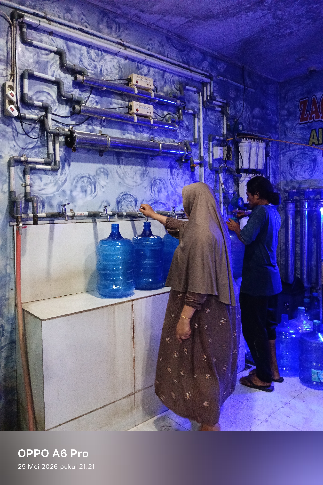 The image shows two people filling large blue water jugs at a water refilling station in Lhokseumawe, Indonesia. A woman in a brown hijab and dress is filling one of the jugs from a tap along a white counter. A man stands next to her, also filling a jug. Behind them is a wall with pipes, filter systems, and other equipment. The lighting is blue, creating a cool and slightly sterile environment. Several filled and unfilled water jugs are placed on the floor. The image was taken with an OPPO A6 Pro on May 25, 2026, at 21:21.
