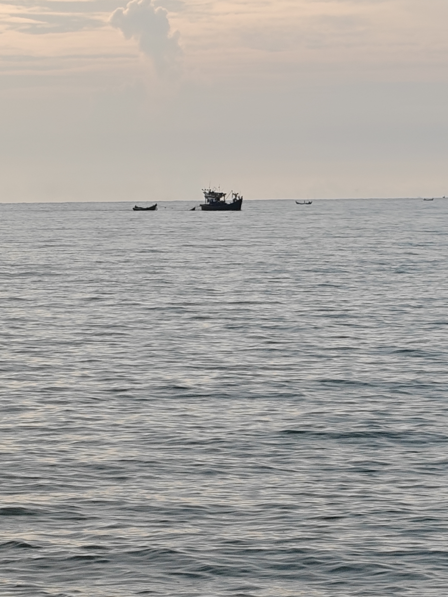 The image shows a calm sea with several boats on the horizon. In the foreground, the water is a deep blue with gentle ripples. The sky above is a pale pink and white, with a large cloud formation in the upper left corner. There are at least three boats visible on the horizon. The largest boat, in the center of the image, appears to be a fishing trawler. To its left, there is a smaller, dark-colored boat. Further to the right, another small boat is visible. The scene appears to be a tranquil morning or evening at sea, possibly near the coast of Lhokseumawe, Indonesia, given the typical maritime activities in the region. The overall impression is one of peaceful solitude.