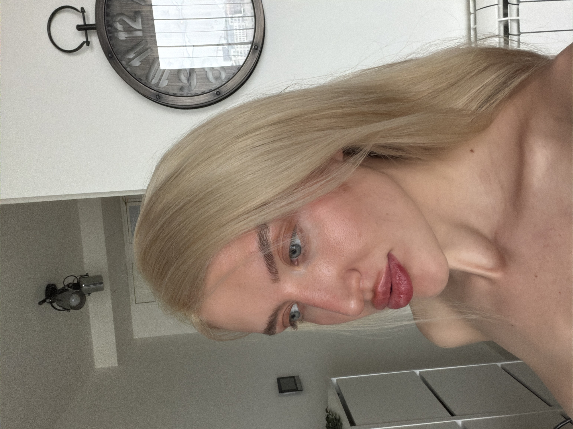 The image captures a close-up of a fair-skinned female with long, straight blonde hair and blue eyes, positioned in an indoor residential setting in Belgrade, Serbia. She faces left, her head slightly lowered, with her bare shoulders and upper chest visible, including a small mole on her right collarbone. Her lips are full and coated with a pink gloss.
The background on the left features a light-colored wall, a partially visible light-toned storage unit with multiple sections, and a small green plant on its top surface. A dark rectangular object is mounted on the wall. Above, a ceiling-mounted light fixture with two dark, adjustable spotlights is visible.
To the right, a large, round, vintage-style wall clock with Roman numerals and a dark metallic frame hangs on a white wall. The clock's glass face reflects an exterior view of multiple distant buildings, indicative of an urban environment. A white radiator with visible fins is present in the lower right portion of the frame.