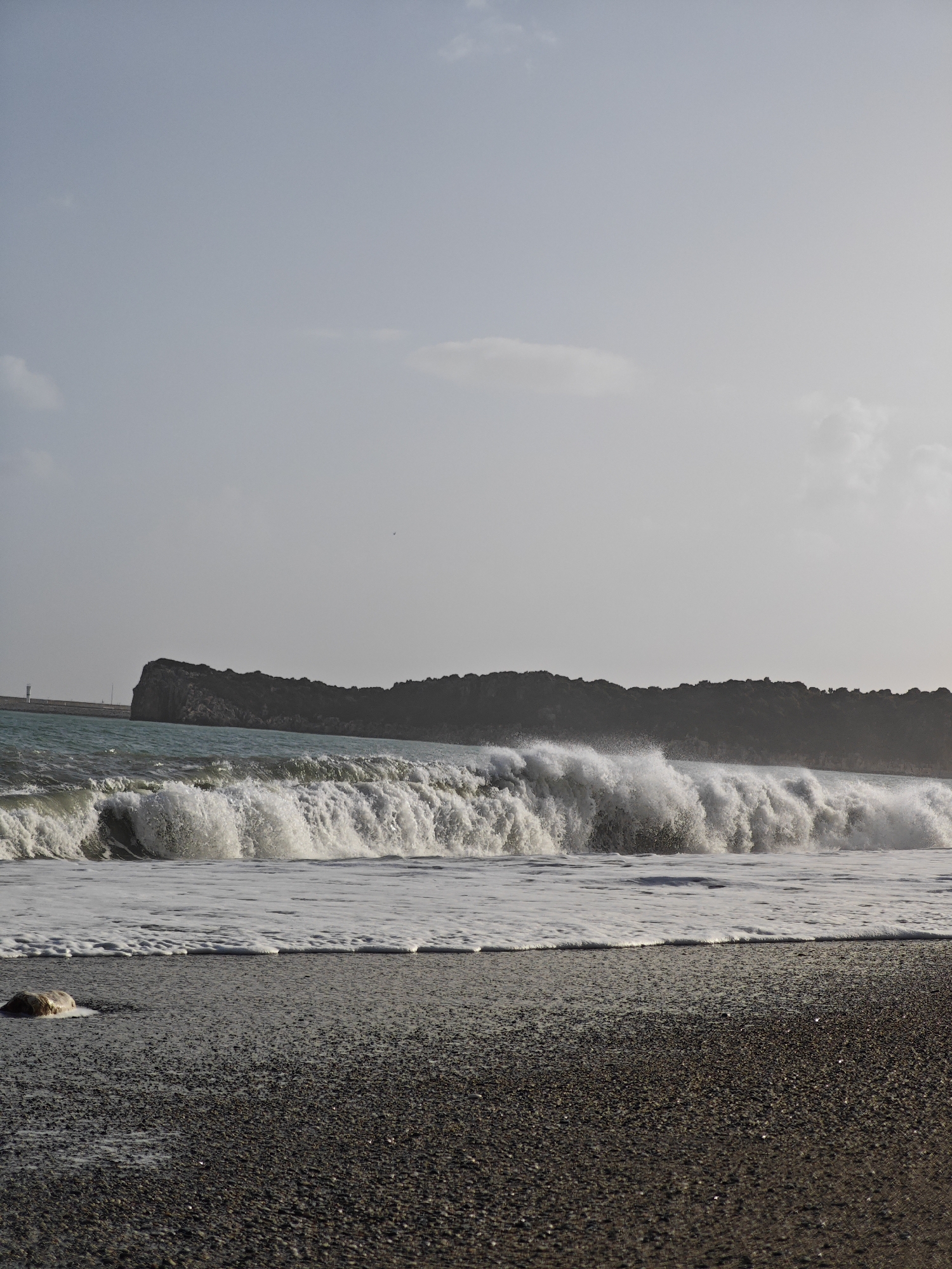 The photograph depicts a scene on a beach with rough, gray-colored sand. A large wave, covered in white foam, is breaking near the shore. In the background, there is a dark, vegetated landmass, and to the left of it, a barely discernible structure resembling a lighthouse or port facility. The sky is overcast with a uniform gray color. The location is most likely a beach in the city of Demre, Turkey, based on the landscape. There are no people visible in the image. A single rock is visible in the lower left corner of the beach.