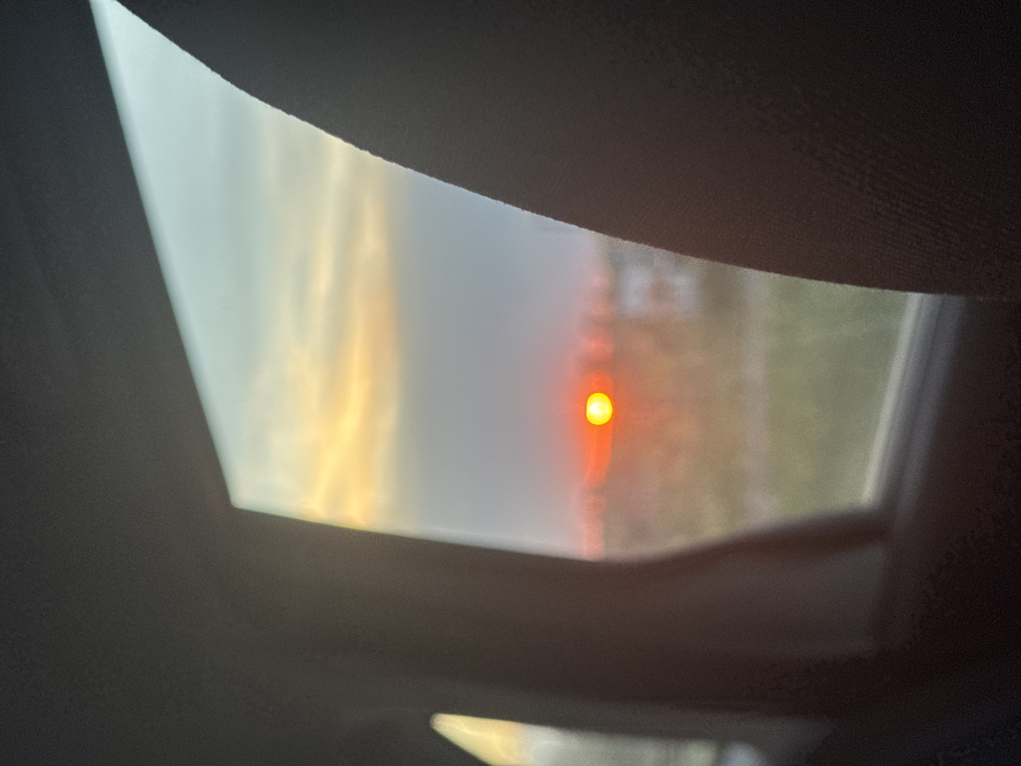 A low-angle, interior view taken through a vehicle's sunroof, partially obscured by the sunroof's interior trim. The sky is visible through the sunroof, appearing hazy or smoky with a low-hanging, intensely red sun. The surrounding environment outside the sunroof is indistinct, with vague indications of trees or other structures. The scene suggests conditions of heavy air pollution or a natural phenomenon such as wildfire smoke. The location is unknown.