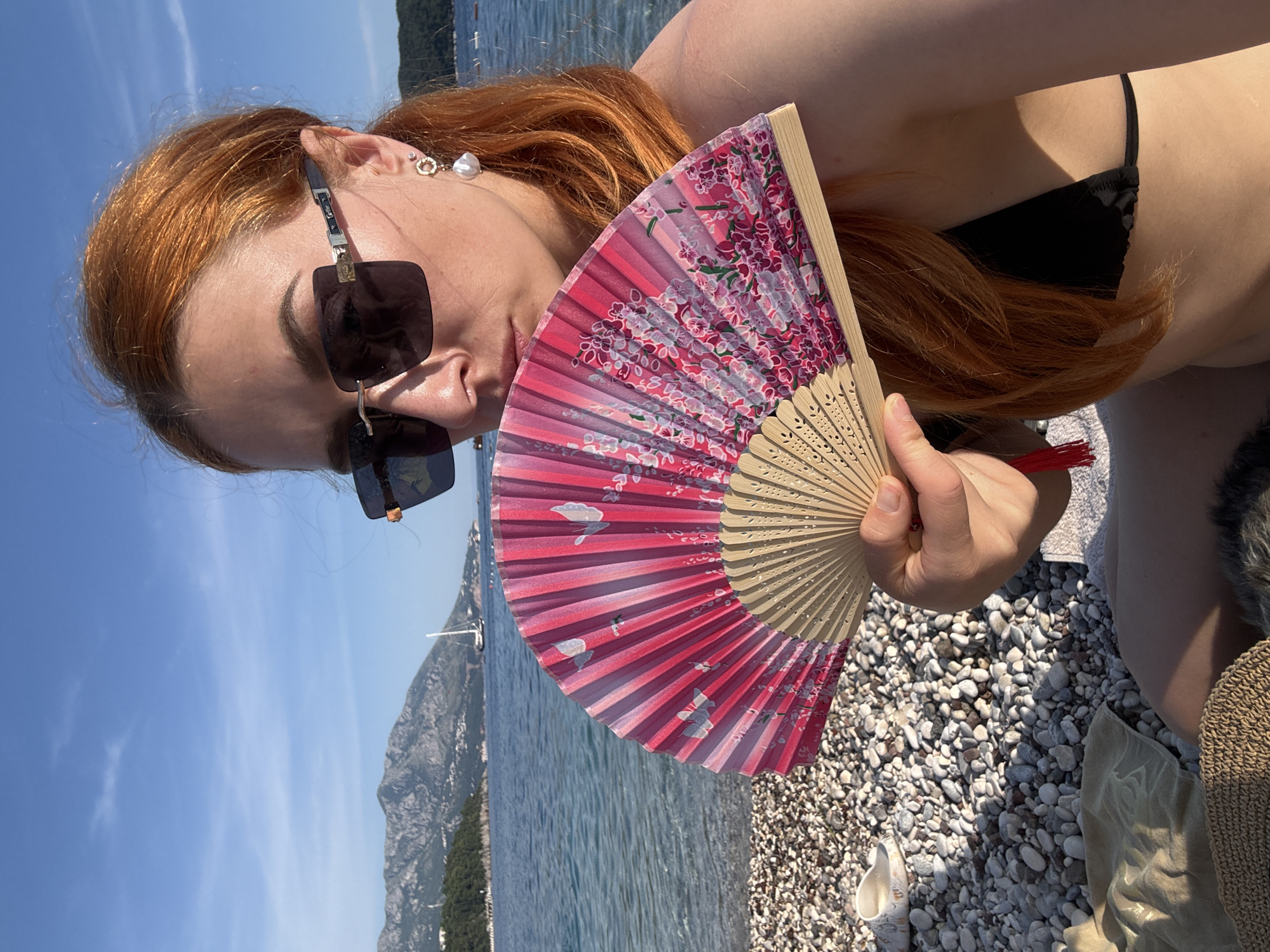 A woman with red hair is holding a pink floral fan in front of her face, while wearing sunglasses and an earring. She is positioned on a pebbled beach, wearing a black bikini. The background features a seascape with mountains, and the sky is blue with light clouds. This image appears to have been taken on a beach in or near Budva, Montenegro, as indicated by the landscape.