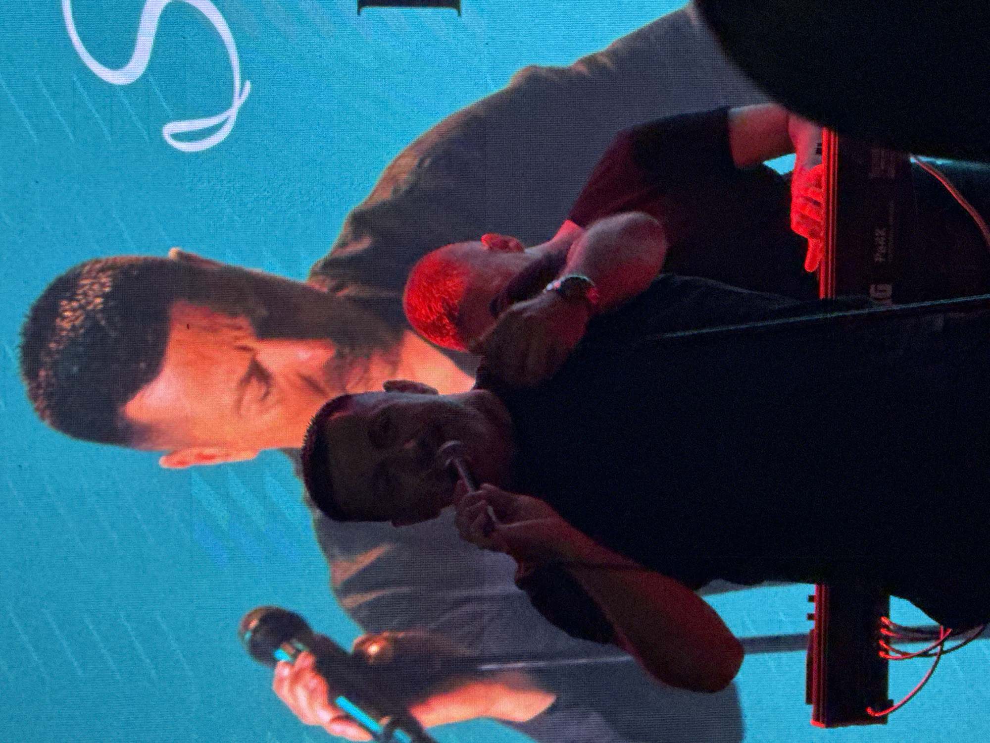 The image shows a close-up of a performer singing into a microphone on a stage, in front of a large screen displaying an image of himself. He is wearing a black t-shirt and is holding the microphone close to his mouth, with his eyes closed or looking down. Behind him, another person with brightly dyed red hair is visible, also holding a microphone. In the foreground, a keyboard or synthesizer is partially visible, with red cables plugged into it. The lighting is dim, with red and blue hues illuminating the scene. The screen behind the performers shows a large, faded image of the lead singer, creating a layered effect. The overall ambiance suggests a live musical performance. The location is likely a music venue in Podgorica, Montenegro, given the context provided in the prompt.