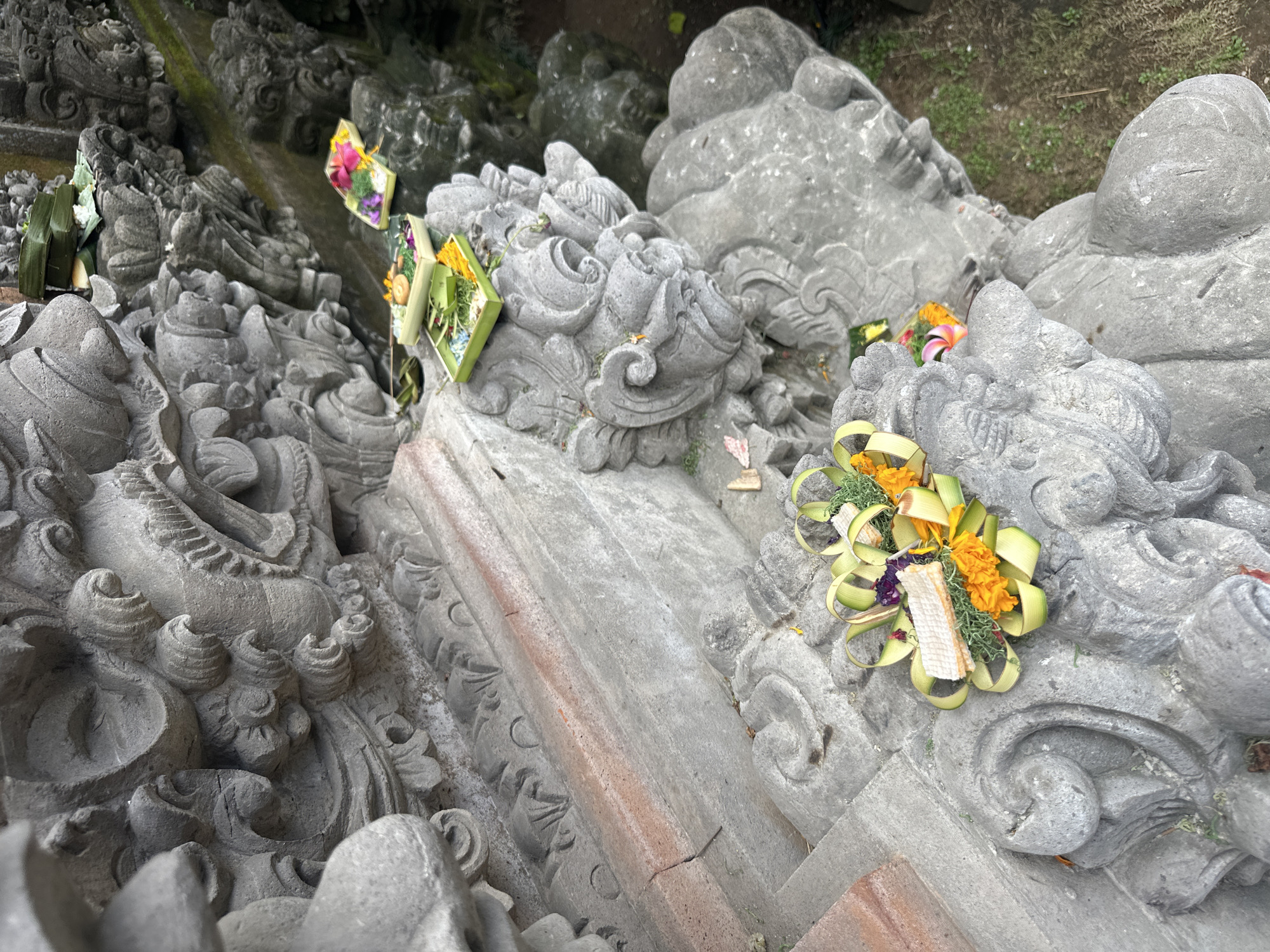An eye-level, medium shot captures a stone carving of a traditional Balinese mythical creature, possibly a Rangda or a similar demon, located in a temple in Ubud District, Indonesia. The carving is weathered and detailed, featuring sharp teeth, elaborate headdresses, and multiple faces. Small offerings, known as *canang sari*, are placed on the stone structures. These offerings consist of woven palm leaves, colorful flowers such as orange marigolds and purple blossoms, and a small white parcel, likely containing rice. In the background, more stone carvings and lush green foliage are visible, suggesting a temple or a sacred site surrounded by nature. The lighting is overcast, casting a soft, diffused light on the scene. The ground beneath is a mix of dirt and dry grass. No people are present in the frame.
