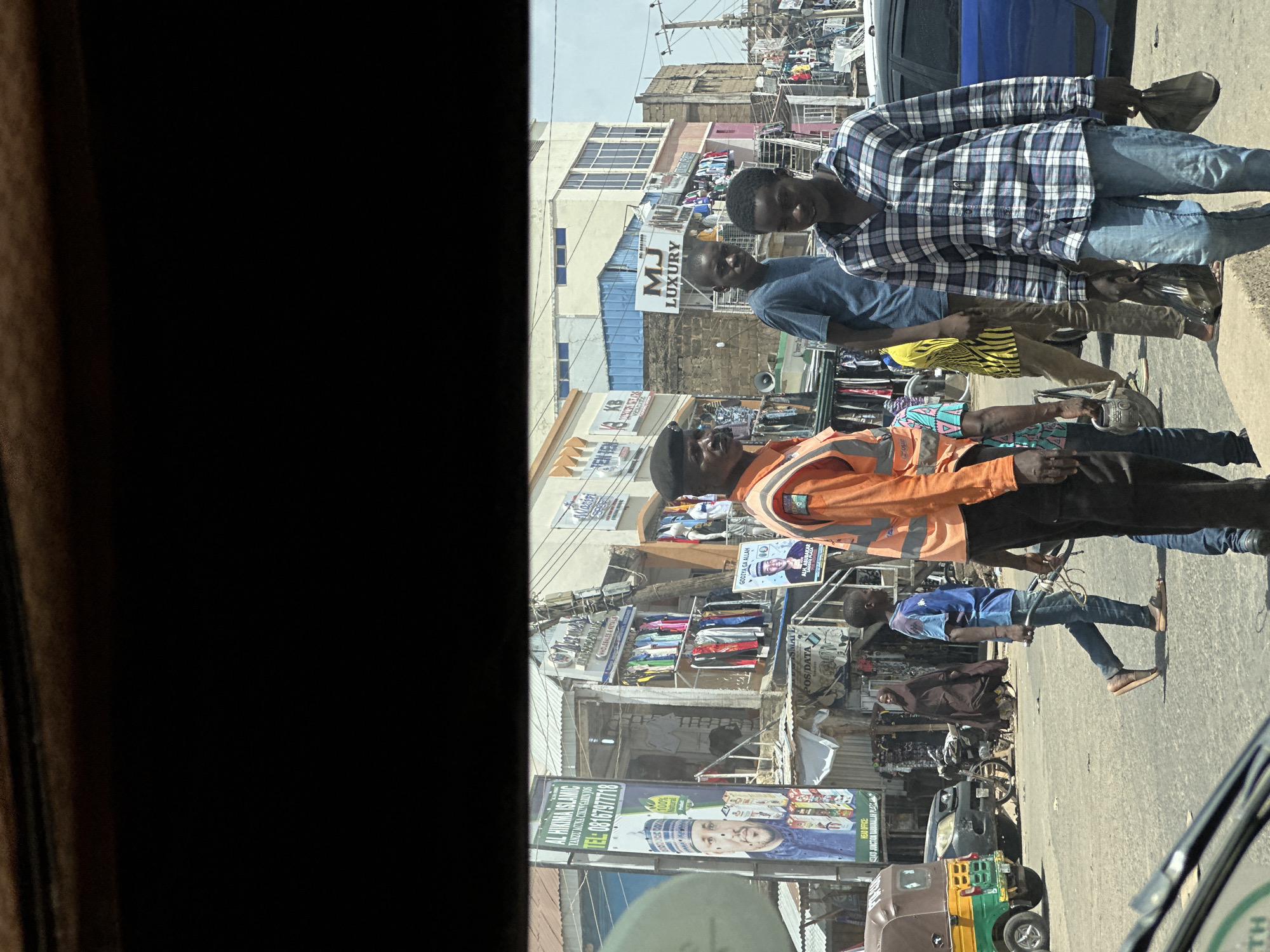 This is a street scene in Jos, Nigeria, featuring a busy marketplace. In the foreground, a man wearing an orange high-visibility vest and a black beret stands to the left. To his right, a young man in a blue and white plaid shirt and jeans walks towards the viewer, carrying two plastic bags. Further back, another young man in a blue t-shirt stands looking towards the viewer. A woman in a dark hijab walks in the background, and another individual in a bright yellow and black patterned shirt is also visible. The environment is characterized by shops with clothing displayed outside on racks, signs advertising businesses like "AL HIKIMA ISLAMIC" and "MJ LUXURY," and various vehicles including a yellow and green auto-rickshaw and a dark SUV. The street is paved, and the overall impression is one of a lively commercial district.