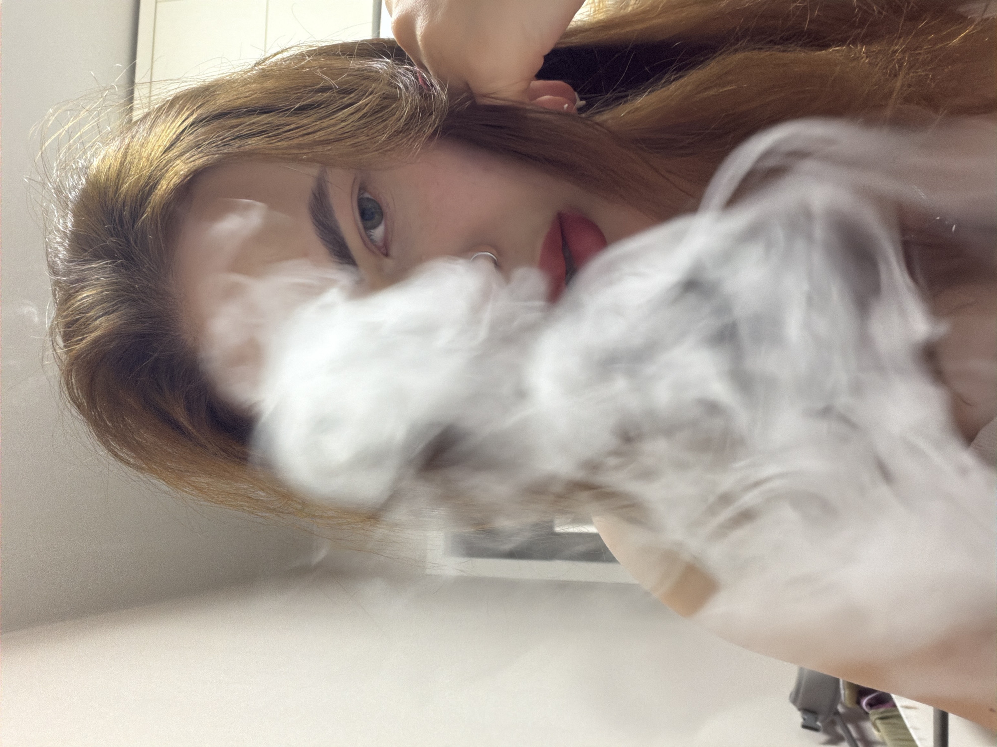 An upward-angled, close-up shot captures a young woman from Krasnodar, Russia, exhaling a dense cloud of white vapor or smoke that partially obscures her face. Her light brown hair is visible, framing her face and catching some light. Her left eye, with a direct gaze, is visible above the smoke, as is her eyebrow. A silver nose ring is prominent on her nostril. Her lips, appearing full and possibly with a light tint, are visible beneath the smoke on the right side of the frame. One of her hands is seen near the top of her head, perhaps holding her hair or resting. The background is softly blurred and consists of light-colored indoor surfaces, suggesting a bright room. The overall impression is intimate and slightly mysterious due to the smoke and the unique perspective. There is no visible text in the image.