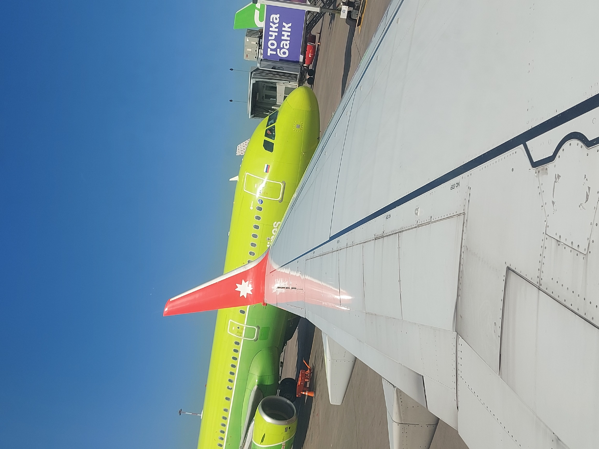 The image shows an airplane wing, presumably from the perspective of a passenger inside the aircraft, with another airplane parked nearby. The parked airplane is painted a bright green and has a red tail with a white star. The "Tochka Bank" sign is visible on the jet bridge. The location is in Saint Petersburg, Russia.