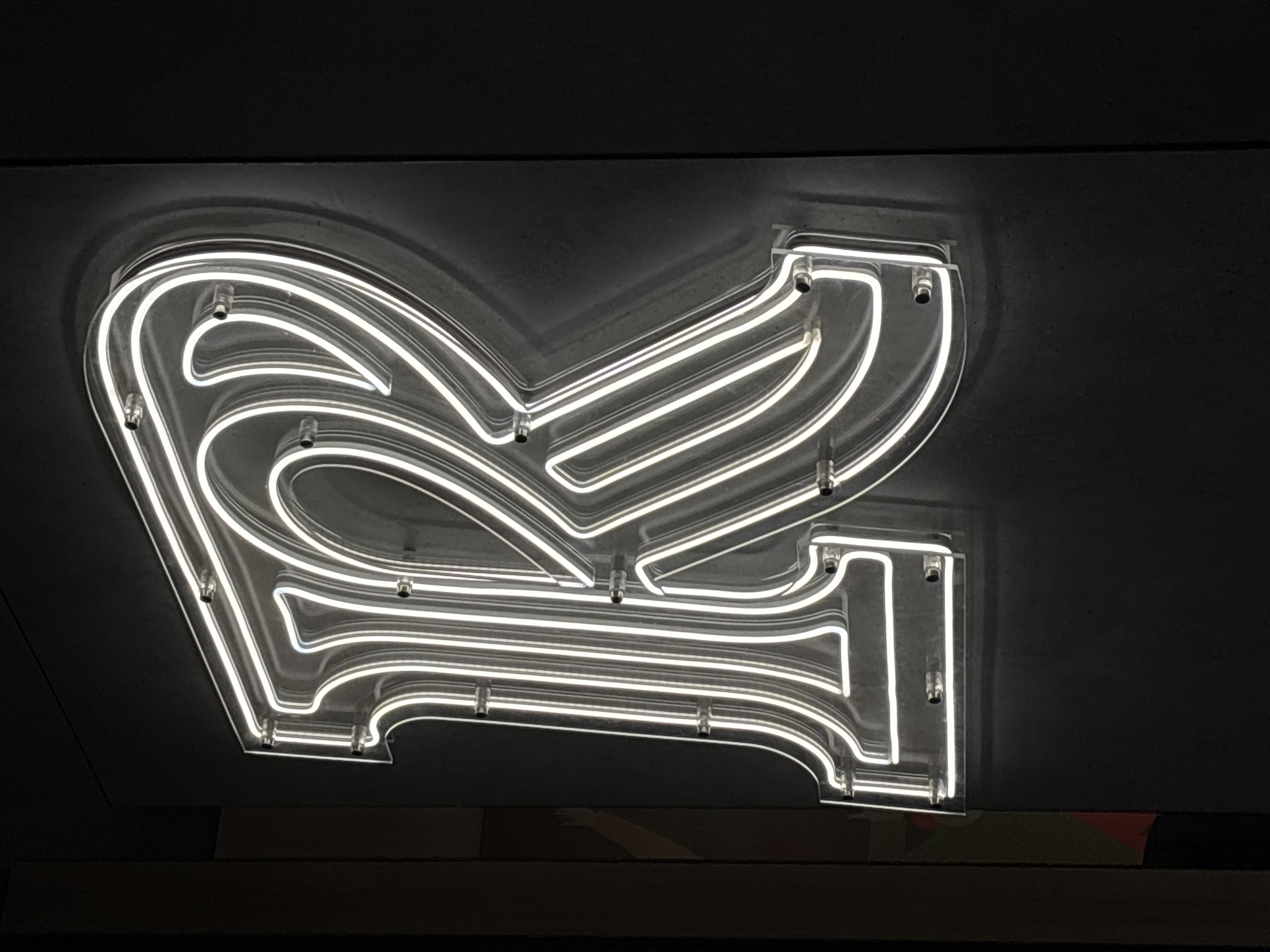 The media file presents a brightly illuminated, stylized white neon sign set against a dark, textured background. The sign is crafted from white LED flex neon tubing, meticulously shaped and mounted onto a clear acrylic panel. Its design appears to be a decorative, flowing capital letter 'R', possibly serving as a logo or an initial. Small, metallic standoffs attach the transparent acrylic backing and the neon tubing to the dark surface, which resembles a wall or ceiling with a matte, possibly concrete-like finish.
The strong contrast between the radiant sign and the deep black background indicates an indoor setting, likely in dim lighting or at night, allowing the sign's glow to stand out prominently. The cool white light emanating from the neon tubing casts a soft illumination on the immediate surrounding dark surface. At the bottom of the frame, beneath the sign, there is a subtle hint of a lighter, horizontal surface, possibly wood. In the bottom right corner, faint abstract colored shapes (red and a lighter tone) are visible, suggesting a design element on a lower structure or surface. This static scene is located in Izhevsk, Russia. The only visible text-like element is the stylized letter 'R' that forms the sign.