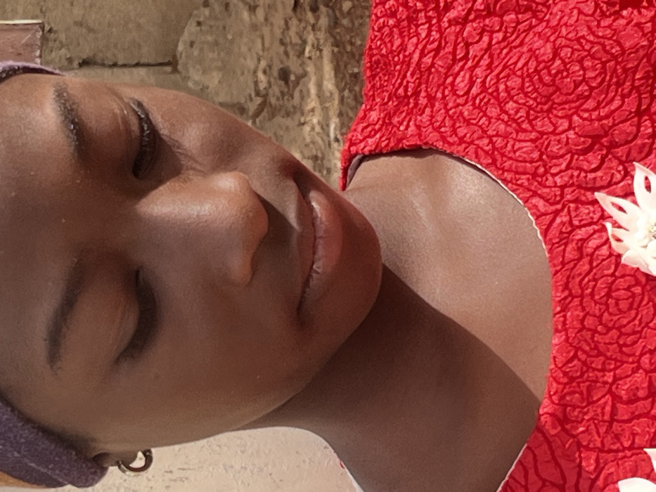 A close-up portrait of an individual, likely female, with dark skin, is depicted outdoors in Jos, Nigeria. The subject's head is angled downwards, with eyes closed or directed towards the ground, and lips gently closed. A purple or dark-colored head covering is visible on the top of the head. A single gold-colored hoop earring is present in the visible left ear. The individual wears a bright red garment featuring a heavily textured, crinkled fabric and a round neckline. A white floral appliqué, with a central pearl-like bead, is positioned on the lower right portion of the garment. The background on the left consists of a plain, light-colored wall, while the right side features a darker, rough-textured wall or stone structure. The scene is illuminated by strong, likely natural, light, creating defined highlights and shadows on the subject and immediate surroundings. No overt actions or interactions are observable.