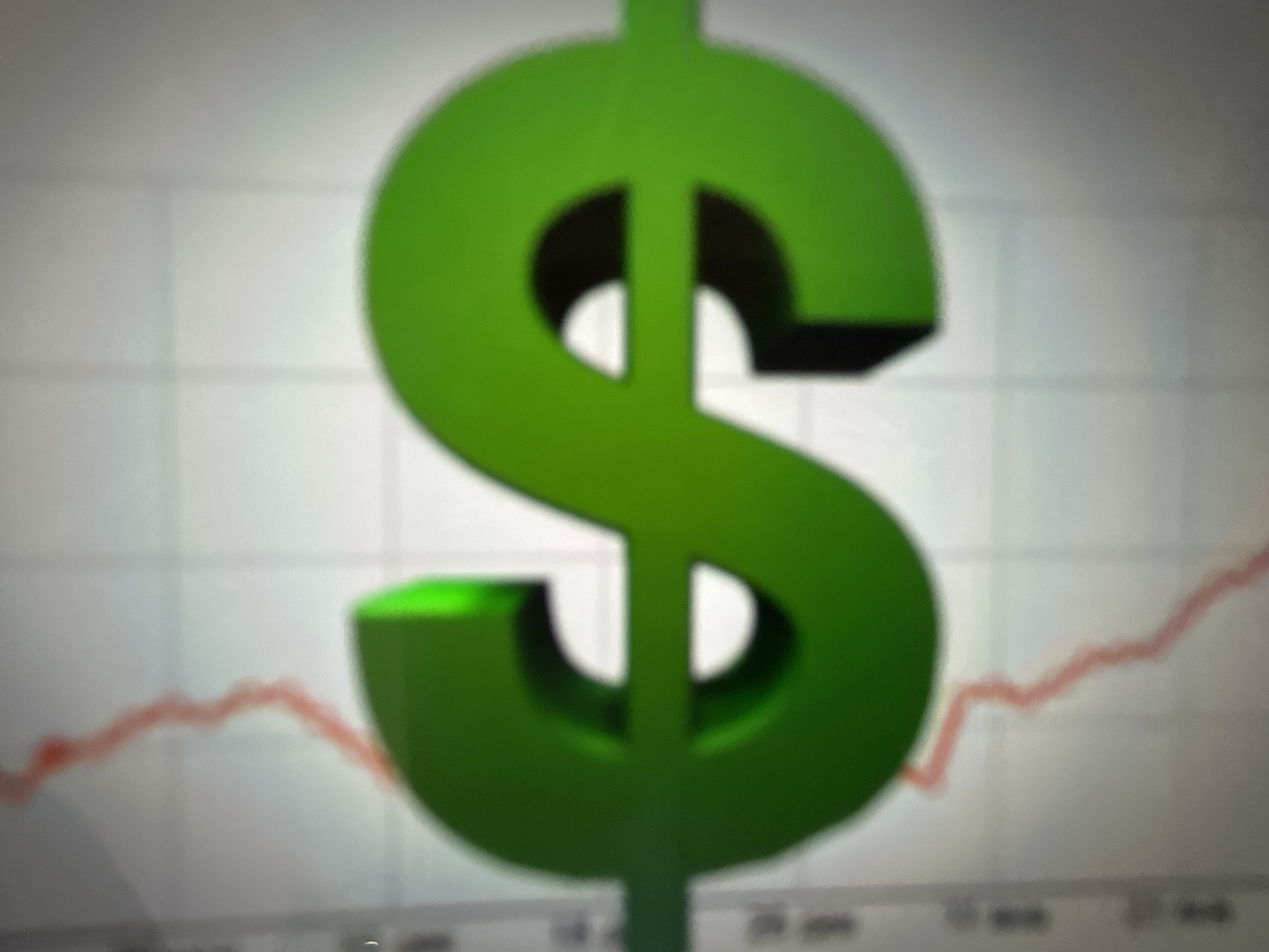 A 3D green dollar sign is superimposed over a blurred financial graph. The graph line is red and appears to be trending upwards, suggesting growth or an increase in value. The background of the graph has a grid pattern, typical of financial charting software. The dollar sign is prominently placed in the center of the image, dominating the foreground. The overall impression is one of financial success or a positive market outlook. There are no people visible in the image, and the setting appears to be digital or abstract, representing financial data. The lighting is bright, and there are no indications of weather or time of day. No discernible text is visible in the image.