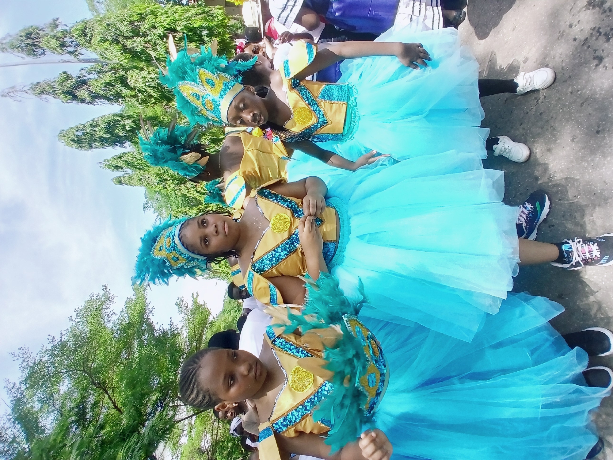 Three young girls are in costume. They are outdoors in Abuja, Nigeria on pavement with trees overhead. The girls wear blue tulle dresses with gold vests and turquoise sequined trim, feathered headdresses, and matching hand-held feathers. The girl in the foreground has braided hair, and the others appear to have closely cropped hairstyles. Adults are visible in the background.