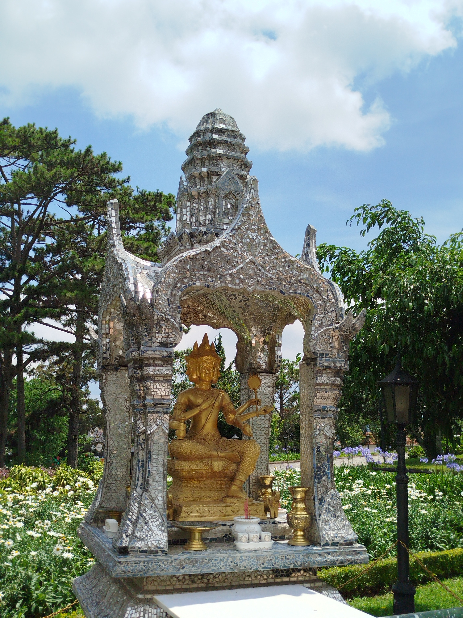 A golden statue of a four-faced deity sits enthroned under a glittering, mosaic-encrusted canopy in the city of Da Lat, Vietnam. The statue is adorned with a conical headdress and appears to hold objects in its multiple hands. The canopy, crafted from mirrored shards, is supported by pillars of similar mosaic work, reflecting light in a dazzling display. The structure rests upon a platform that also features the mosaic technique. In front of the statue, there are ceremonial items, including incense and small containers. The backdrop features a lush garden with a variety of plants and flowers, adding a natural element to the scene. To the right, a black lamppost stands amidst the foliage. Tall trees and a bright blue sky with fluffy white clouds complete the composition.