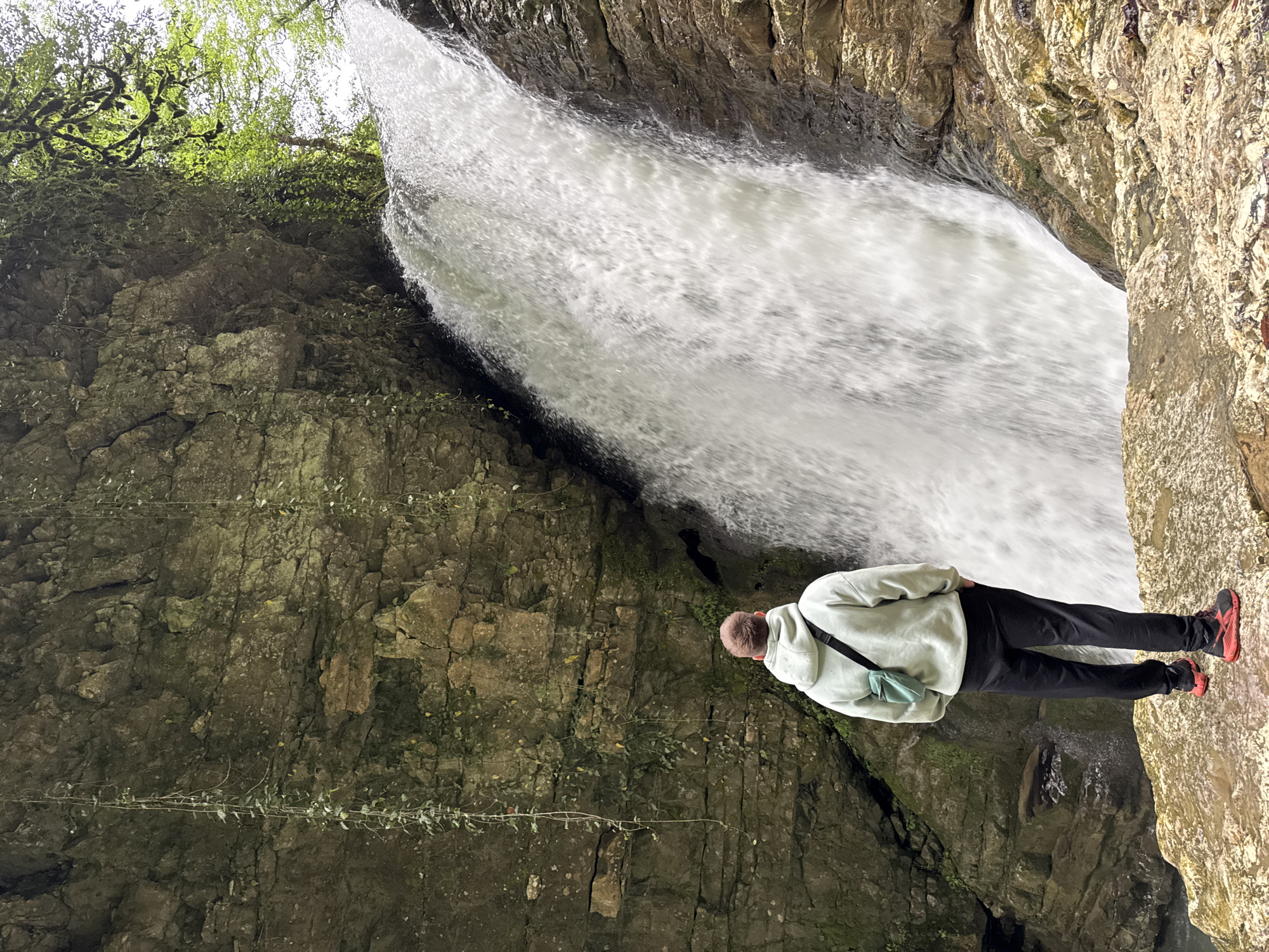The image depicts a person standing near a waterfall. The person is wearing a light-colored hoodie, dark pants, and red shoes. They are facing towards the waterfall, hands on their hips, with a small bag slung across their shoulder. The waterfall is situated within a rocky cliffside, with green foliage visible at the top. The rock formations surrounding the waterfall are textured and weathered. The location is Tsabal, Abkhazia.