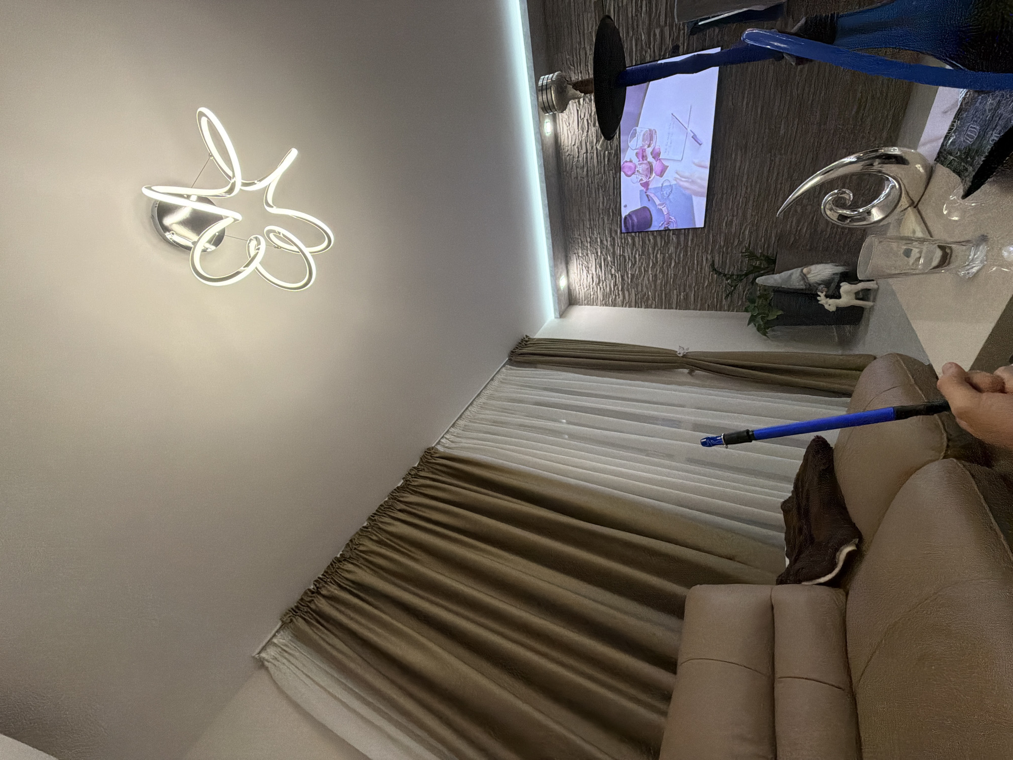 The image displays a modern living room or lounge area, observed from a slightly elevated angle. On the light-colored ceiling, a distinctive modern chandelier with abstract, flowing white LED light elements is prominently featured. A linear LED strip light also illuminates the ceiling along the junction with the wall.
A flat-screen television is mounted on a textured, dark grey wall, displaying an image that appears to be a close-up of hands arranging pink rose petals and wine glasses, possibly a still from a video or a romantic-themed advertisement. Below the television, a shelf or console holds various decorative objects: a shiny, silver, swirling abstract sculpture, a small garden gnome figurine, a miniature white deer figurine, and a potted green plant. Partially visible above and to the right of the TV, there is a metallic base with a dark bowl and blue tubing, characteristic of a hookah.
To the right of the room, a large window is covered with curtains, comprising inner sheer white panels and outer opaque, pleated brown or golden-beige drapes. In the foreground, part of a beige or light brown upholstered sofa or armchair is visible, with a dark brown cloth draped over its armrest. A person's hand is visible on the right, holding the blue hose of the hookah, which has a black mouthpiece or handle. The room is brightly illuminated by the artificial light from the chandelier and LED strip, suggesting it is evening or night. No discernible text is visible in the image.
