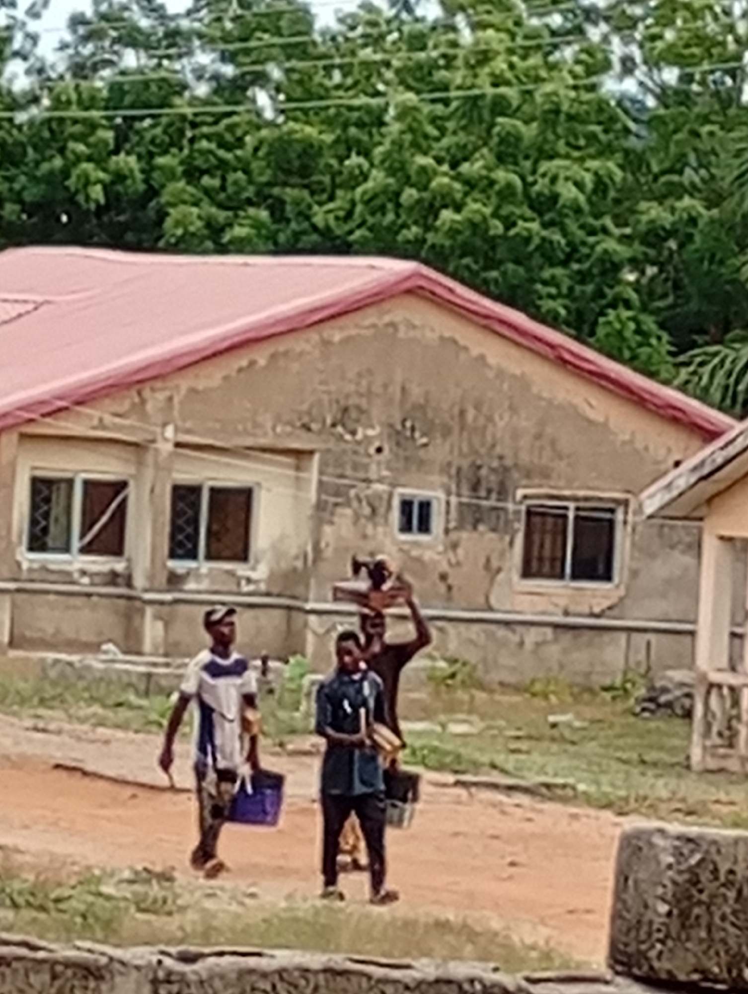 The image shows three individuals walking in front of a building with a pink roof. One person is carrying items on their head, another is holding a blue container, and the third person is walking in the middle. The building has multiple windows, and there are trees in the background. The location appears to be in a residential area. The city and specific neighborhood are not identifiable from the image.