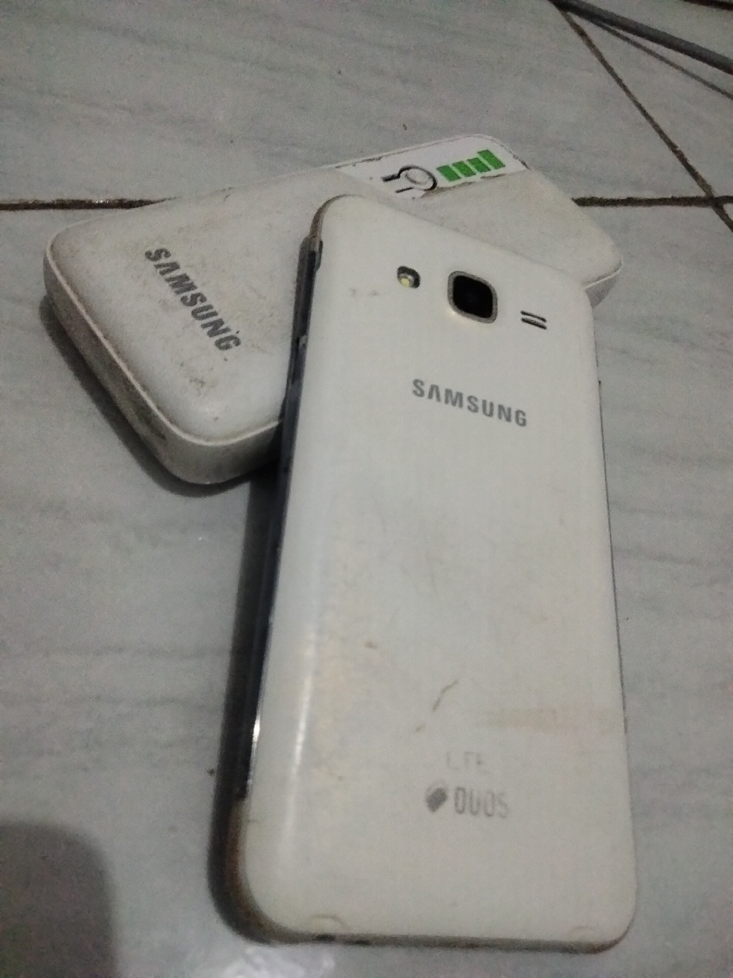The image shows a white Samsung mobile phone and a white Samsung power bank resting on a tiled surface. The power bank is positioned behind and slightly to the left of the phone. The phone is oriented vertically, with its back facing the camera. The power bank is also oriented horizontally.
On the power bank, the word "SAMSUNG" is visible in black text. There is also a graphic of a battery with several green bars, indicating a charge level, and a power symbol.
On the back of the phone, the "SAMSUNG" logo is printed in black text near the top. Below that, there are two small speaker grilles. Further down, the text "LTE" is visible, followed by a diamond-shaped icon and the word "DUOS" in black text, indicating it's a dual-SIM phone.
The objects appear to be used and are covered in a layer of dust or dirt, suggesting they are not new. The lighting is moderate, and there are some shadows cast, but no specific time of day can be determined. The tiled surface suggests an indoor setting, possibly a floor or a table. The background is blurred, with some indistinct lines that might be part of furniture or cables. There are no people or animals in the image.
The overall scene is a still life composition of two electronic devices. The visible text identifies the brands and some features of the products.