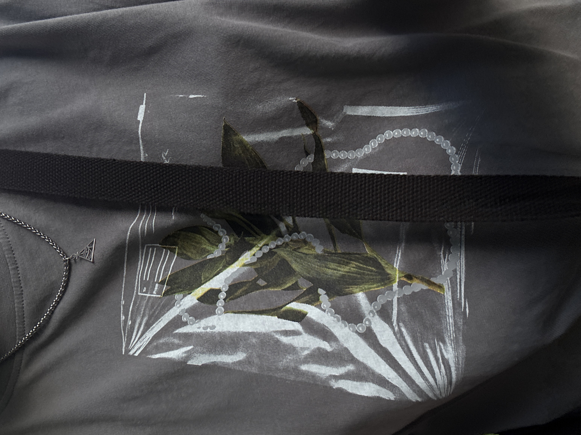 The image shows a close-up of a person wearing a dark grey t-shirt with a graphic print of a plant encased in plastic wrap, adorned with pearls. A silver chain necklace with an eye-shaped pendant is visible around the neck. A dark strap, likely from a bag, crosses the t-shirt diagonally. The background is indistinct and appears to be an indoor setting with neutral lighting. The image does not provide any information about the location within the city of Saint Petersburg, Russia.
