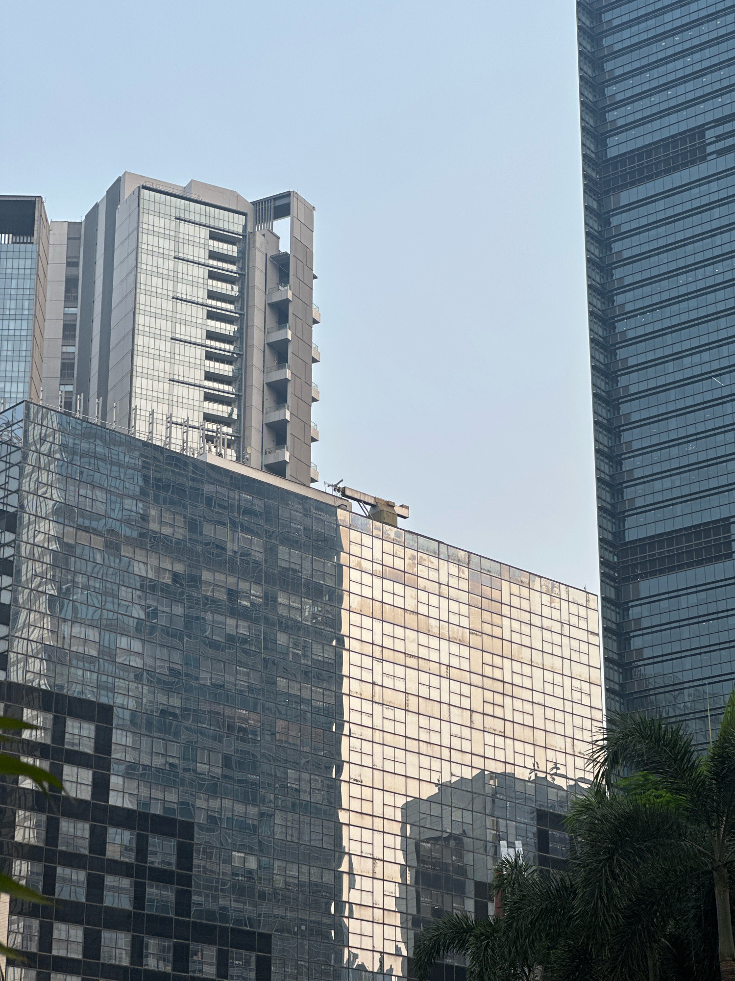 The image shows a low-angle view of modern skyscrapers in South Jakarta, Indonesia, under a clear sky. The buildings are characterized by their glass facades, reflecting the surrounding environment and the sky. One building on the right is dark blue-tinted glass, while another in the center features a more complex design with visible balconies and architectural details. A lower building in the foreground has a reflective glass facade that is partially golden in hue, possibly due to the angle of the sun. Palm trees are visible in the lower right corner, adding a tropical element to the urban landscape. There are no people or discernible activity visible in the image, presenting a static architectural scene.