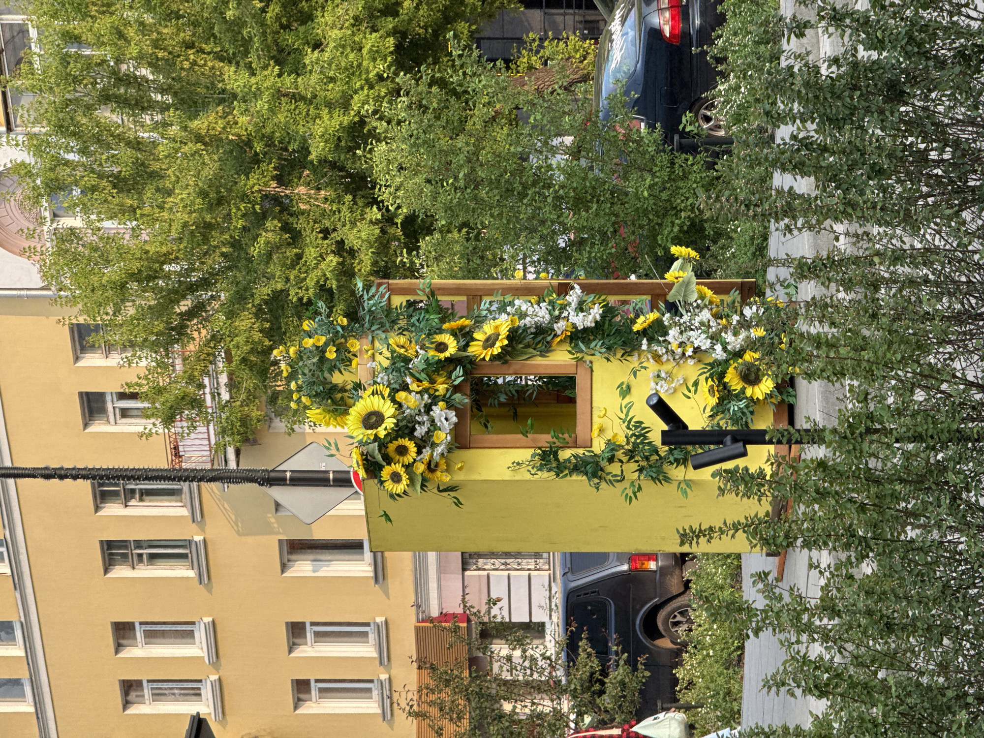 The image depicts an outdoor scene in the city of Krasnoyarsk, Russia. In the foreground, a dense hedge of green bushes lines a paved walkway. Behind the hedge, a bright yellow, booth-like structure is decorated with artificial sunflowers and white flowers. The structure has a wooden-framed window opening. A black pole with two spotlights is attached to the front of the booth. To the left of the yellow structure, a dark-colored SUV is parked on the street. In the background, a building with a yellow facade and multiple windows stands. Lush green trees are visible behind the building. The lighting suggests it is daytime, likely late afternoon or early evening due to the warm tones of the sunlight.