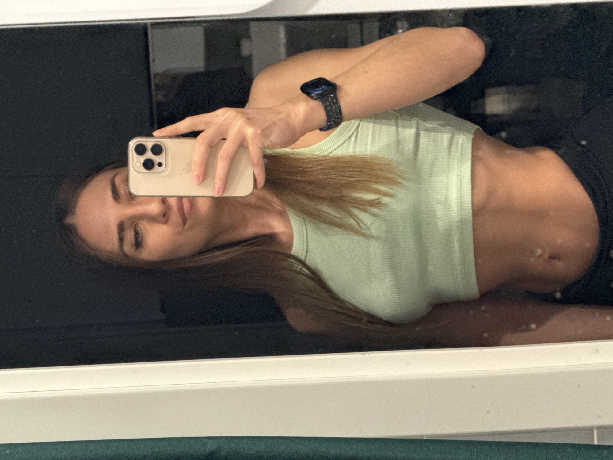 A young woman is taking a selfie in a mirror. She is wearing a light green tank top, black leggings, and a smartwatch with a dark-colored band. She is holding a smartphone with a triple-lens camera in her left hand. The reflection in the mirror shows a black surface behind her, possibly part of the bathroom decor. The mirror is framed with a white border. The location is not discernible from the image.
