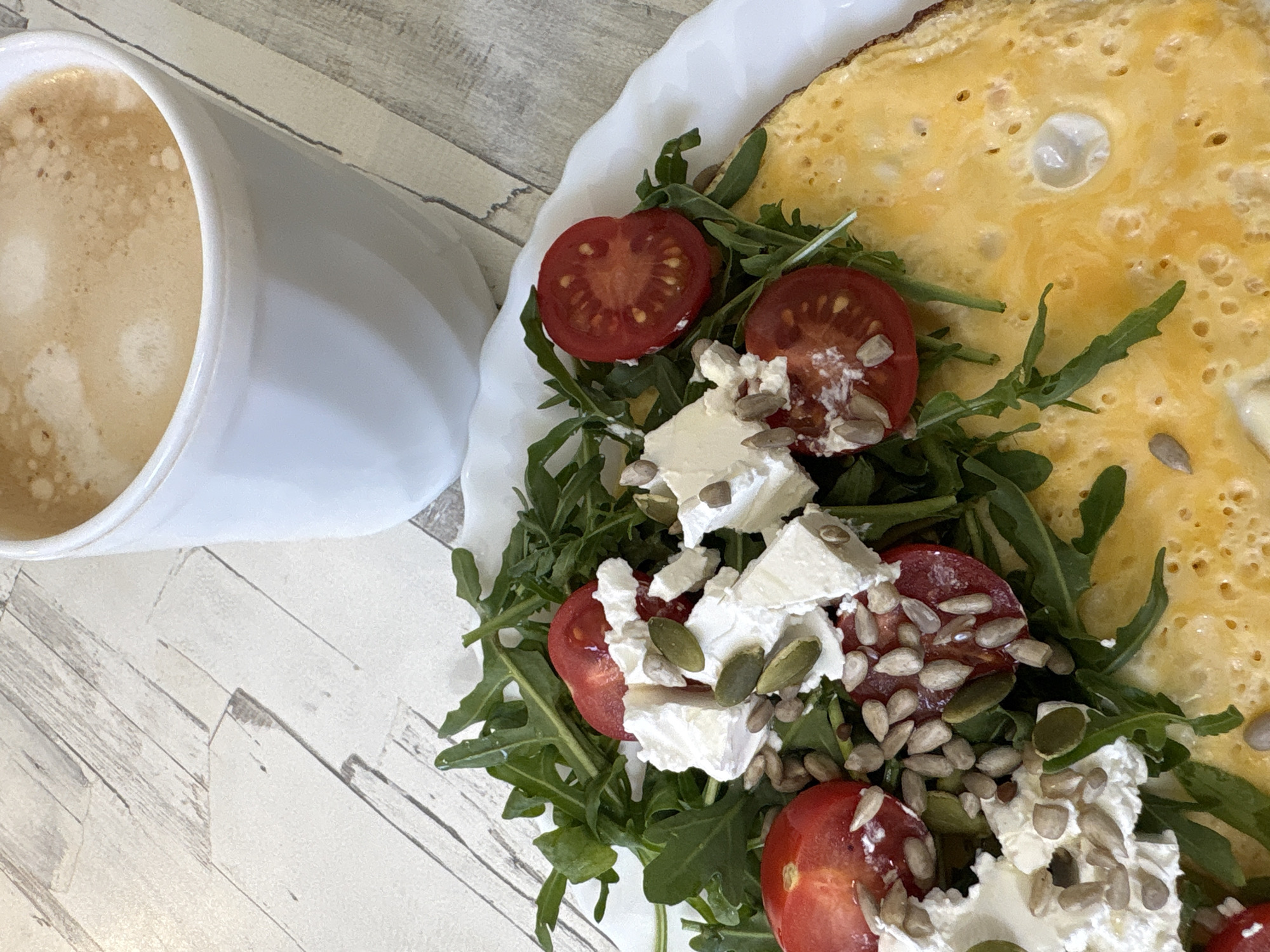 A high-angle shot depicts a breakfast scene, likely indoors on a light, wood-patterned surface. On the left is a white mug filled with a latte. To the right, a white plate holds an omelet, topped with a salad. The salad consists of arugula, halved cherry tomatoes, cubed white cheese (possibly feta), and a mix of pumpkin and sunflower seeds. There is no indication that the location is Chaikovsky, Russia, or any other specific location. No people are visible. The scene focuses on the breakfast elements.