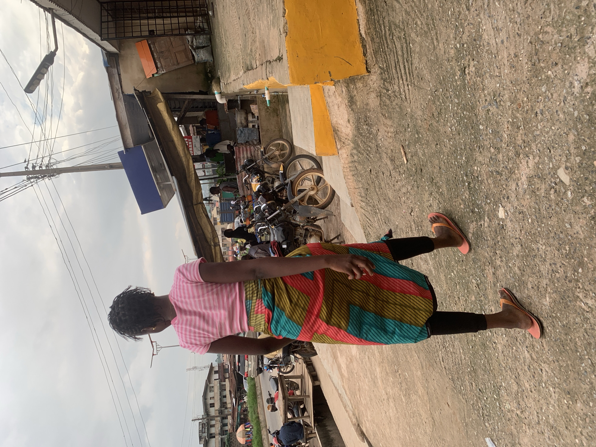 The image depicts a street scene in Shagamu, Nigeria, with a young woman walking away from the camera. She is wearing a pink horizontally striped t-shirt and a vibrantly colored, patterned wrap skirt with stripes of red, yellow, green, and blue. Black leggings are visible beneath the skirt, and she is wearing orange flip-flops. The background shows a mix of buildings, a street with passing vehicles and motorcycles, and what appears to be a market or roadside stalls. Several motorcycles are parked on the right side of the image, and a yellow painted concrete structure is prominent in the foreground. Overhead, power lines and a street light are visible against a cloudy sky.
