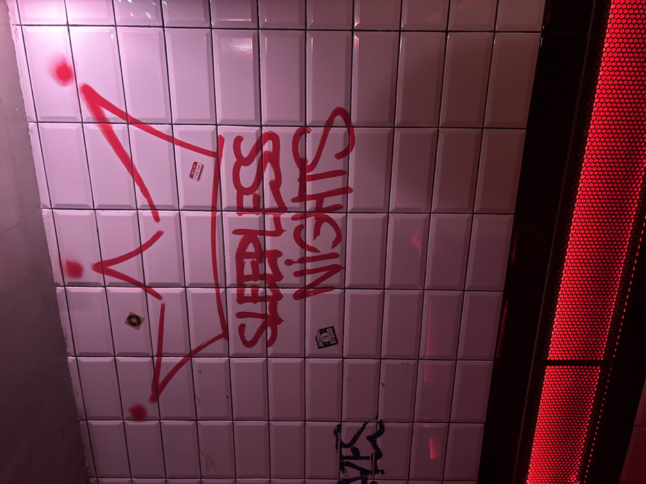 A white tiled wall in the Madrid Metro system, Madrid, Spain, is illuminated primarily by pink and red lighting. The wall consists of numerous rectangular, beveled white ceramic tiles arranged in horizontal rows. Centered on the wall is a red spray-painted crown design, beneath which the phrase "SLEEPLESS NIGHTS" is spray-painted in red capital letters. To the left, partial black graffiti spelling "YFS" is visible. Several stickers are affixed to the tiles: a small, circular black and yellow sticker with an unclear symbol is located within the left portion of the crown. A white rectangular sticker with red text, reading "HELLO I'M A BAD IDEA," is present near the right peak of the crown. A black rectangular sticker displaying "ARCANGEL STUDIO" in white text is positioned below the "SLEEPLESS NIGHTS" graffiti. A smaller, dark sticker is also visible below the "YFS" graffiti. Along the base of the wall, a horizontal black structure houses a bright, perforated red light panel, which emits a strong red glow upwards across the lower section of the tiles. No individuals are present in the scene.