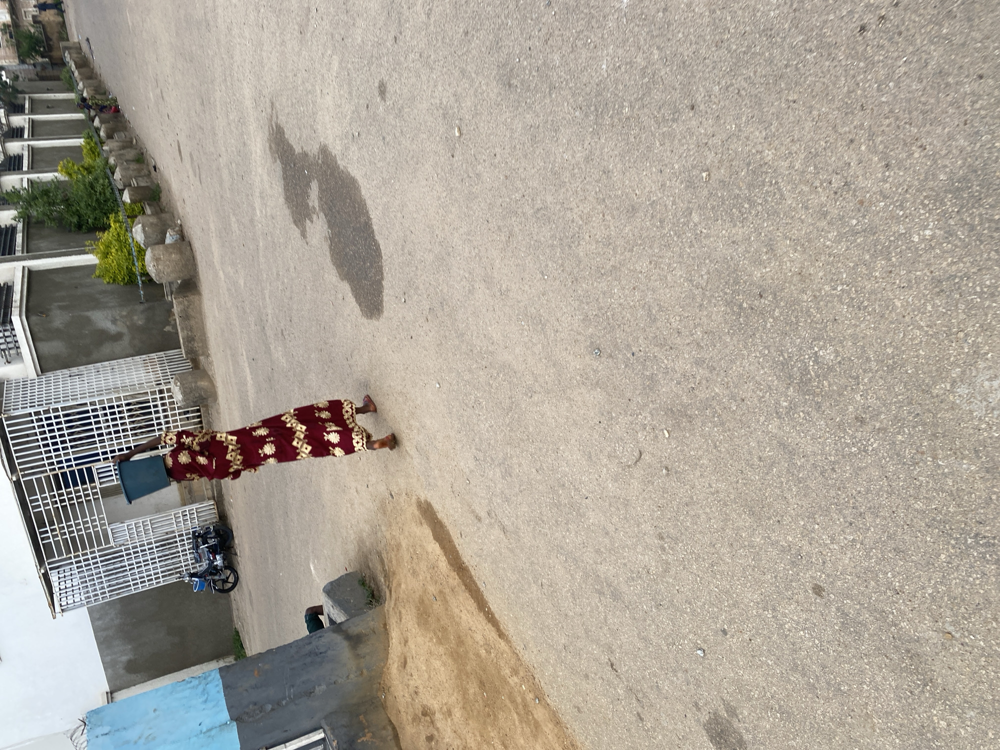 The image shows a person standing on an asphalt road in what appears to be an urban or semi-urban environment in Jos, Nigeria. The person is wearing a long, patterned dress and is holding a light blue object, possibly a container, in front of them. They are standing near a white metal gate or fence, and a motorcycle is parked behind the gate.
To the left of the person, there are buildings with white exteriors and grey sections. Green foliage, including trees and bushes, is visible in front of these buildings. The road surface is grey asphalt with some visible stains and debris. The scene suggests it is daytime, and the lighting indicates fair weather. The perspective of the image is from a slightly elevated viewpoint looking down at the scene. No text is visible in the image.