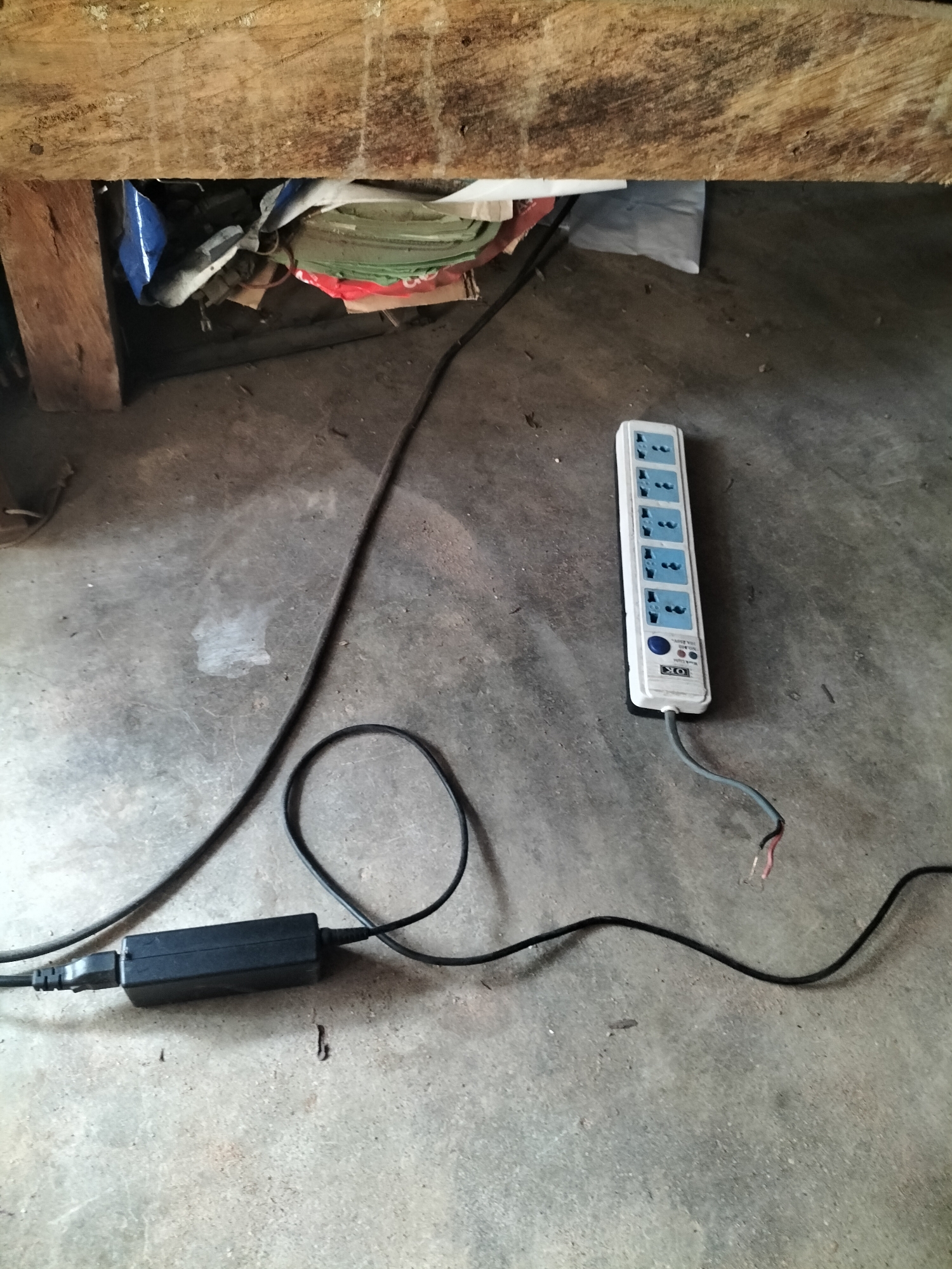 The image shows a power strip with multiple outlets and a black power adapter lying on a concrete floor. The power strip is white with blue outlets and a blue button, and it has a cord with exposed wires. The power adapter is also black. In the background, there are some items stacked under a wooden surface. The scene appears to be in a dimly lit, possibly storage area. The provided context places this scene in Jalingo, Nigeria.