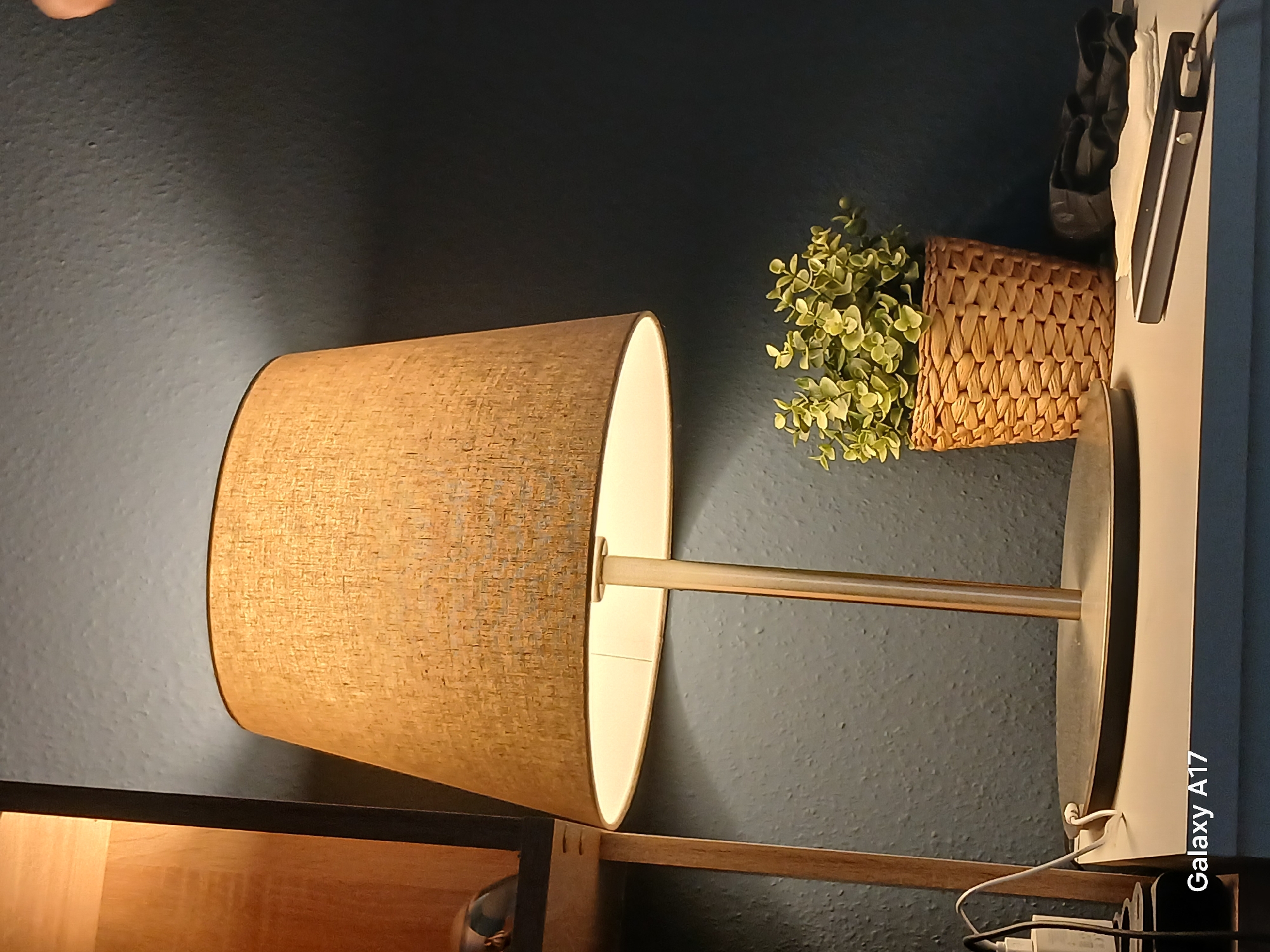 The image shows a lamp with a textured lampshade, casting a warm glow. To the right of the lamp is a small, woven basket containing a faux plant with light green leaves. A white power strip is visible near the lamp's base. The background is a dark teal or blue wall. The image appears to have been taken indoors, and "Galaxy A17" is visible in the lower right corner. No people are visible. There is no information to suggest the location is Yekaterinburg, Russia.