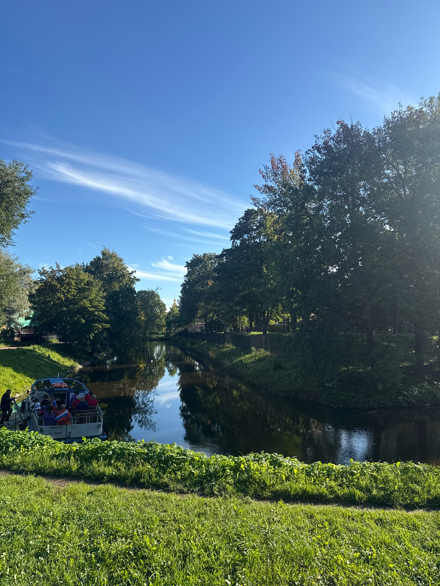 This image captures a scenic canal in Saint Petersburg, Russia, on a bright, sunny day. The sky is a vibrant blue with wispy white clouds streaking across it. The canal itself is calm and reflects the surrounding greenery and the sky. On the left side of the canal, a tour boat with several passengers is docked, appearing to be either boarding or disembarking. The boat has the Cyrillic text "ФИЕСТА" and license plate "03 09-26" visible on its stern. Lush green grass and trees line the banks of the canal, creating a peaceful and picturesque atmosphere. The trees are full of leaves, suggesting it is either late spring or summer. The overall impression is one of a pleasant day for outdoor activities in a beautiful urban setting.
