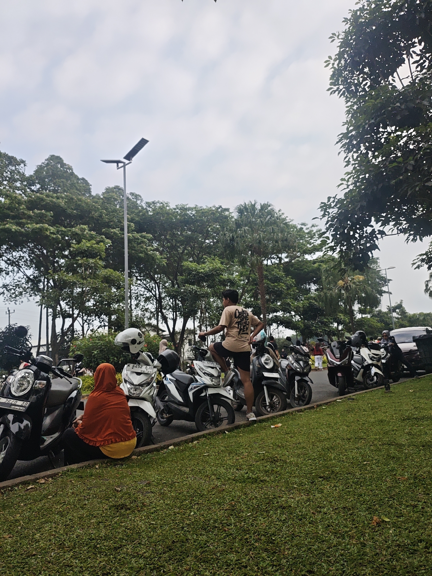 In the city of Bacin, Indonesia, a group of motorbikes is parked on the side of a grassy area. The scene is set outdoors under an overcast sky. In the foreground, several motorbikes are lined up, with one person in an orange hijab and yellow skirt sitting on the curb in front of a white motorbike. To the left, a black motorbike is parked, and further back, more motorbikes are visible. A man in a light brown t-shirt and dark shorts is standing on one of the motorbikes, looking away from the camera. Behind the motorbikes, a row of trees forms a backdrop, with a tall street light visible in the distance. Some people are also seen in the background, and a portion of a car is visible on the far right. The ground is covered in well-maintained green grass.