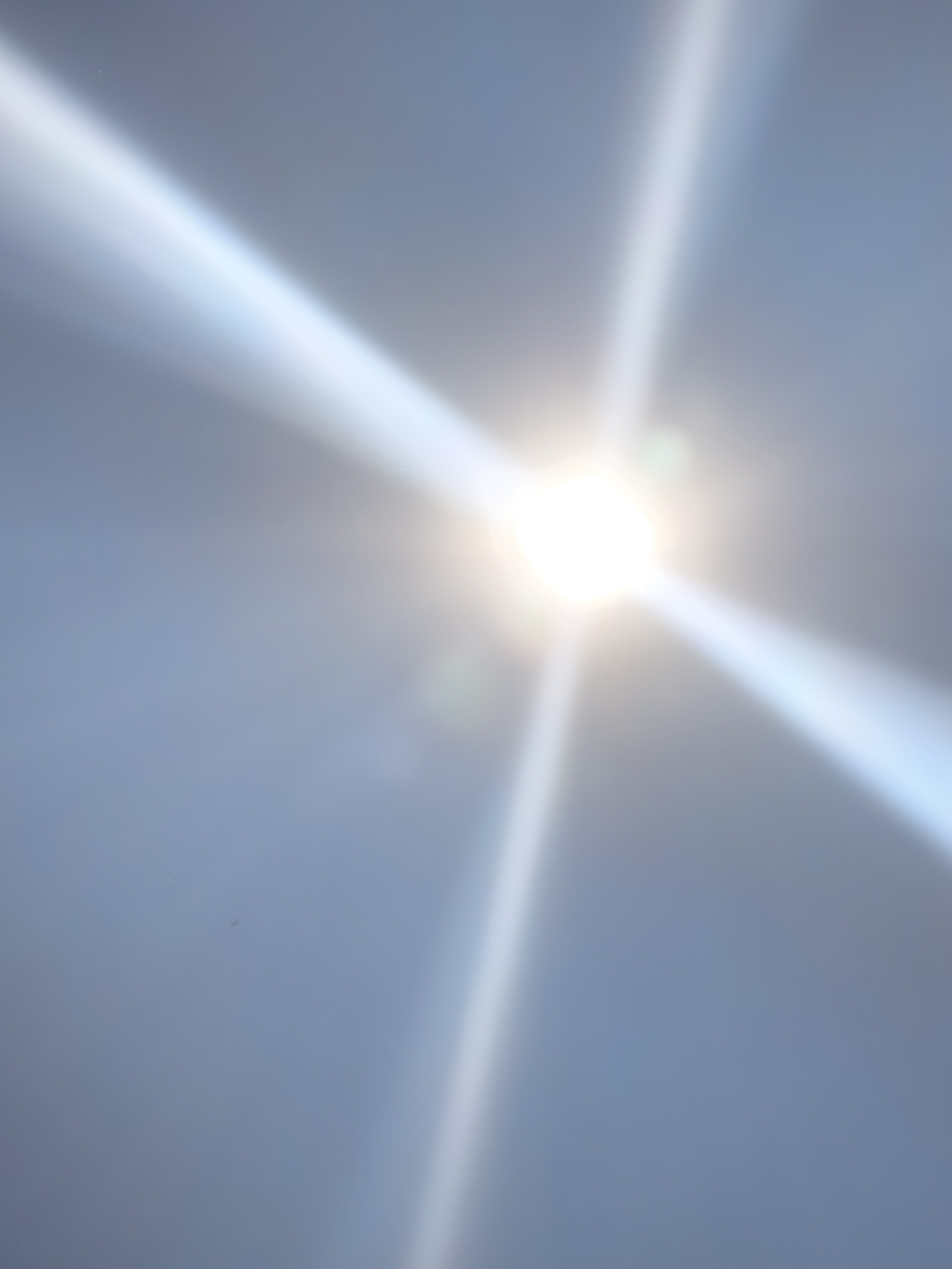 This media file is an abstract photograph of the sun with lens flare, set against a clear sky. The sun appears as a bright, white light at the center of the image, with prominent rays of light extending outwards in a cross shape. The sky is a pale blue, gradually becoming darker towards the edges of the frame.
There are no people or specific objects visible in the scene. The overall impression is one of brightness and expansive sky. The light rays create a dramatic and ethereal effect, suggesting a powerful light source. A very small, dark speck, possibly a distant bird or insect, is visible in the upper left quadrant of the image. Another small, faint green flare is visible near the sun.
The time of day is likely daytime, given the presence and brightness of the sun. The weather appears clear and sunny, with no clouds obscuring the sky. There are no specific location cues, making the setting indeterminate. The image does not convey any discernible emotions; it is purely a visual representation of light and sky. There is no visible text in the media file.
